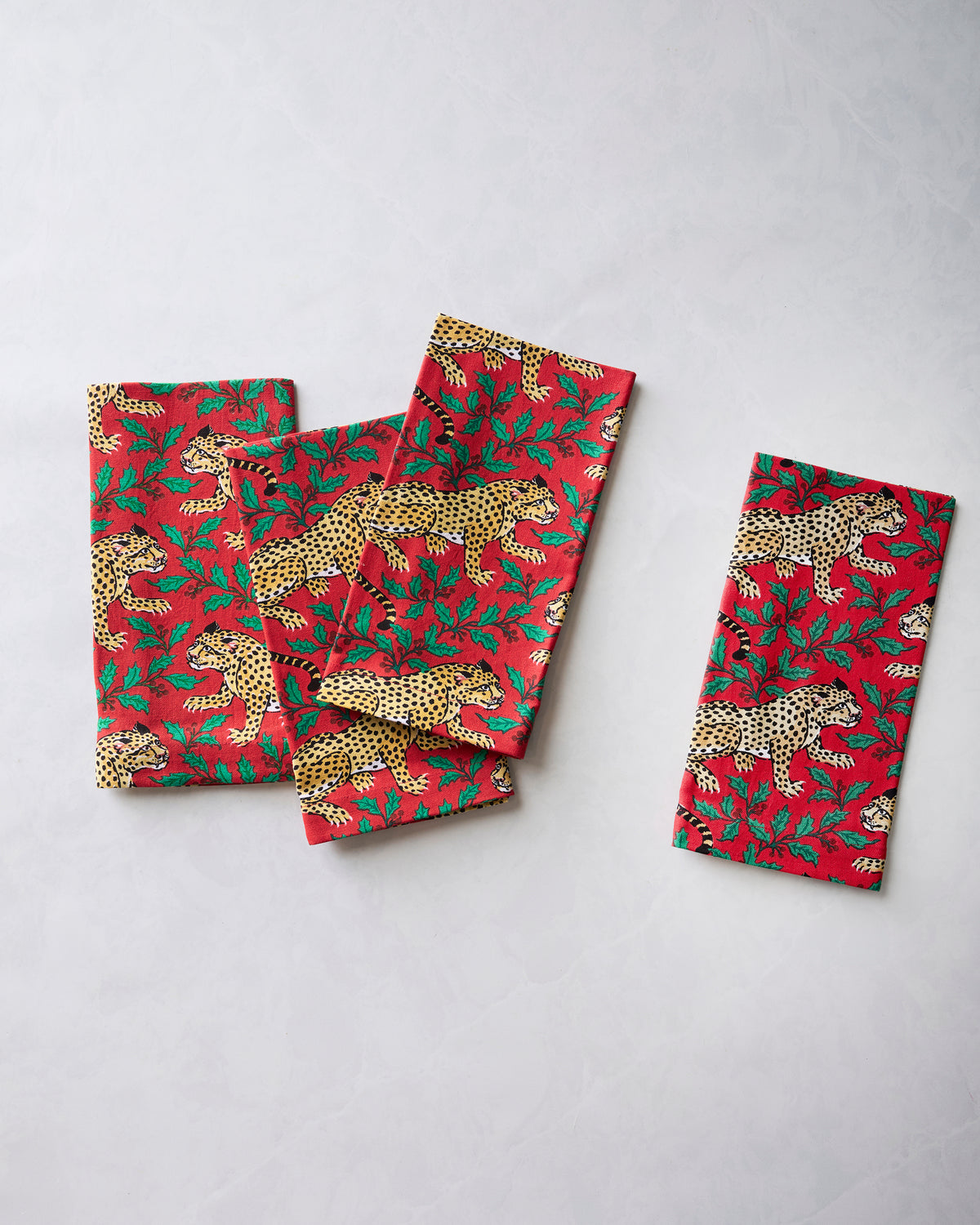 Four ruby dinner napkins with leopard and holly print arranged in a stack, with one napkin displayed separately.