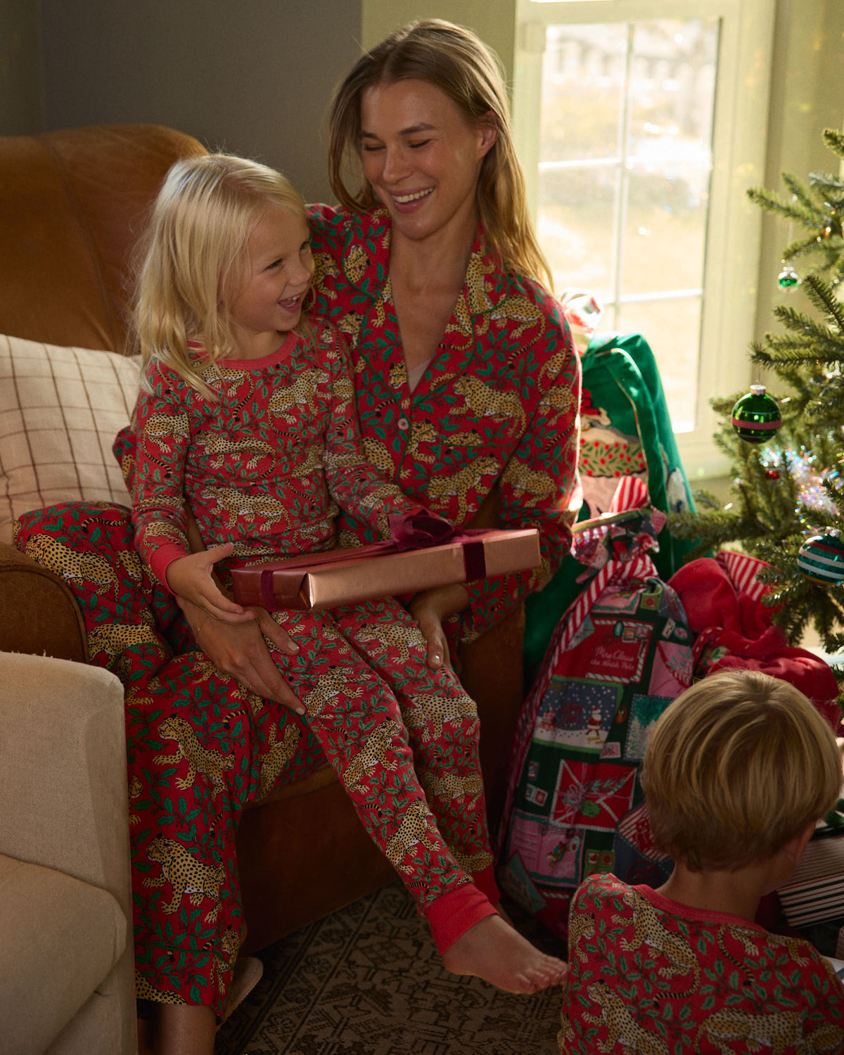 Adult and child wearing matching red leopard-and-holly pajamas sit together near a Christmas tree with wrapped gifts.