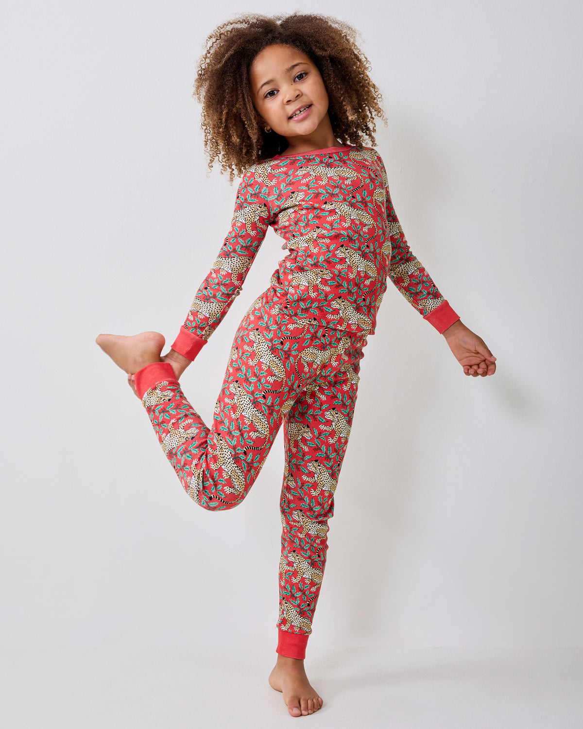 Child modeling a red long-sleeve pajama set with spotted leopards and holly leaves while standing.