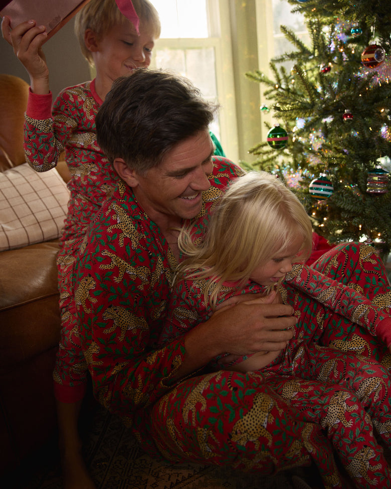 Adult holding a child in matching red leopard-and-holly pajamas near a decorated Christmas tree.