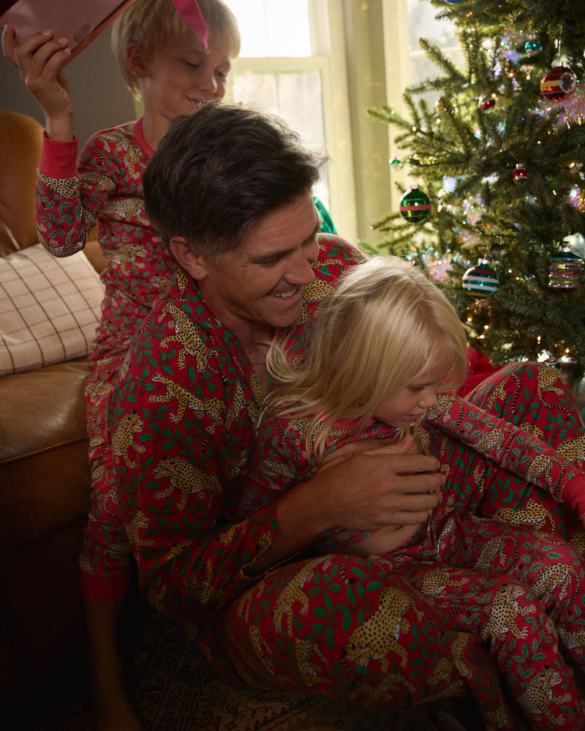 Adult holding a child in matching red leopard-and-holly pajamas near a decorated Christmas tree.