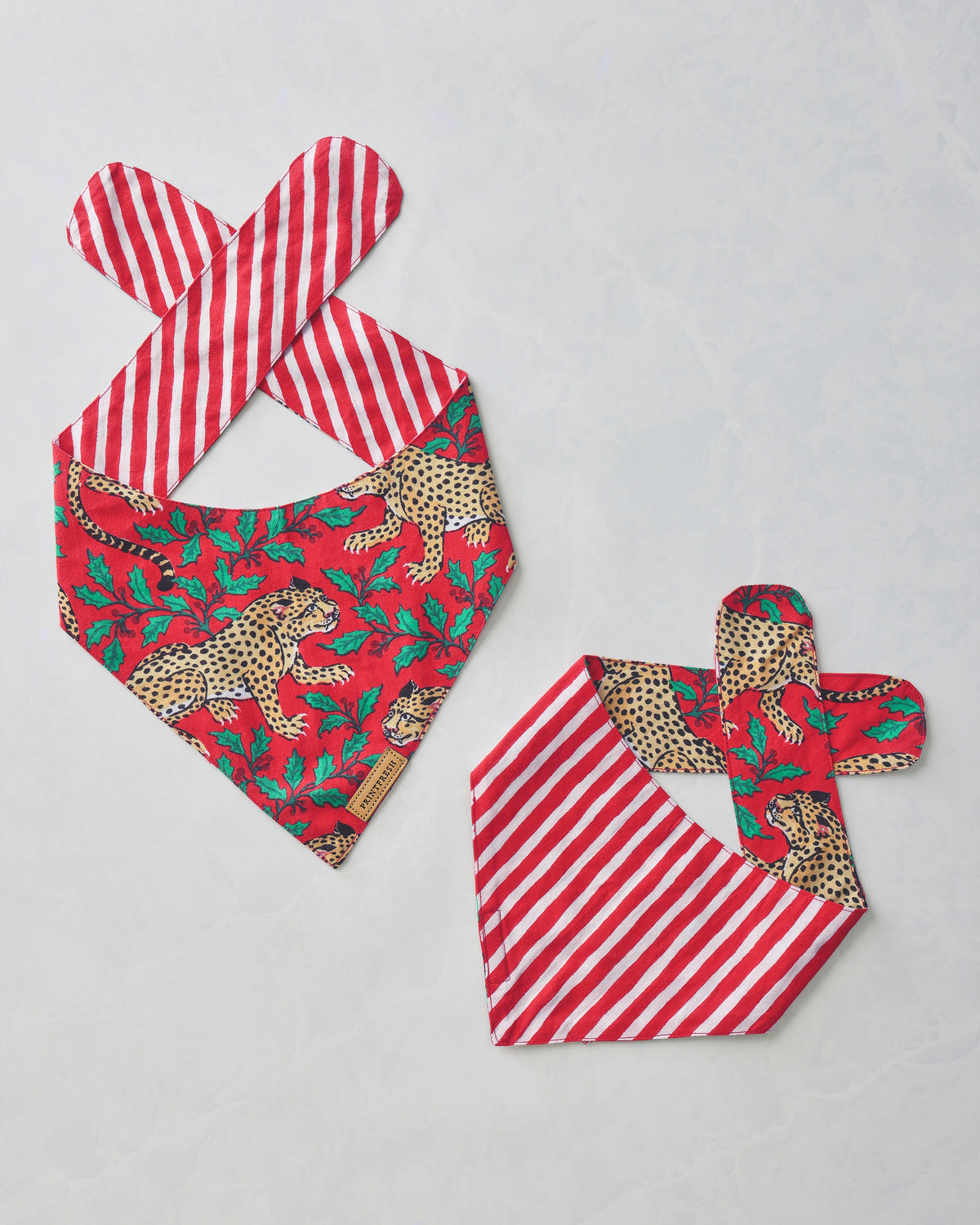 Two Ruby Winter Holly Bagheera pet bandanas shown front and reverse, with leopard and holly print on one side and red-and-white stripes on the other.
