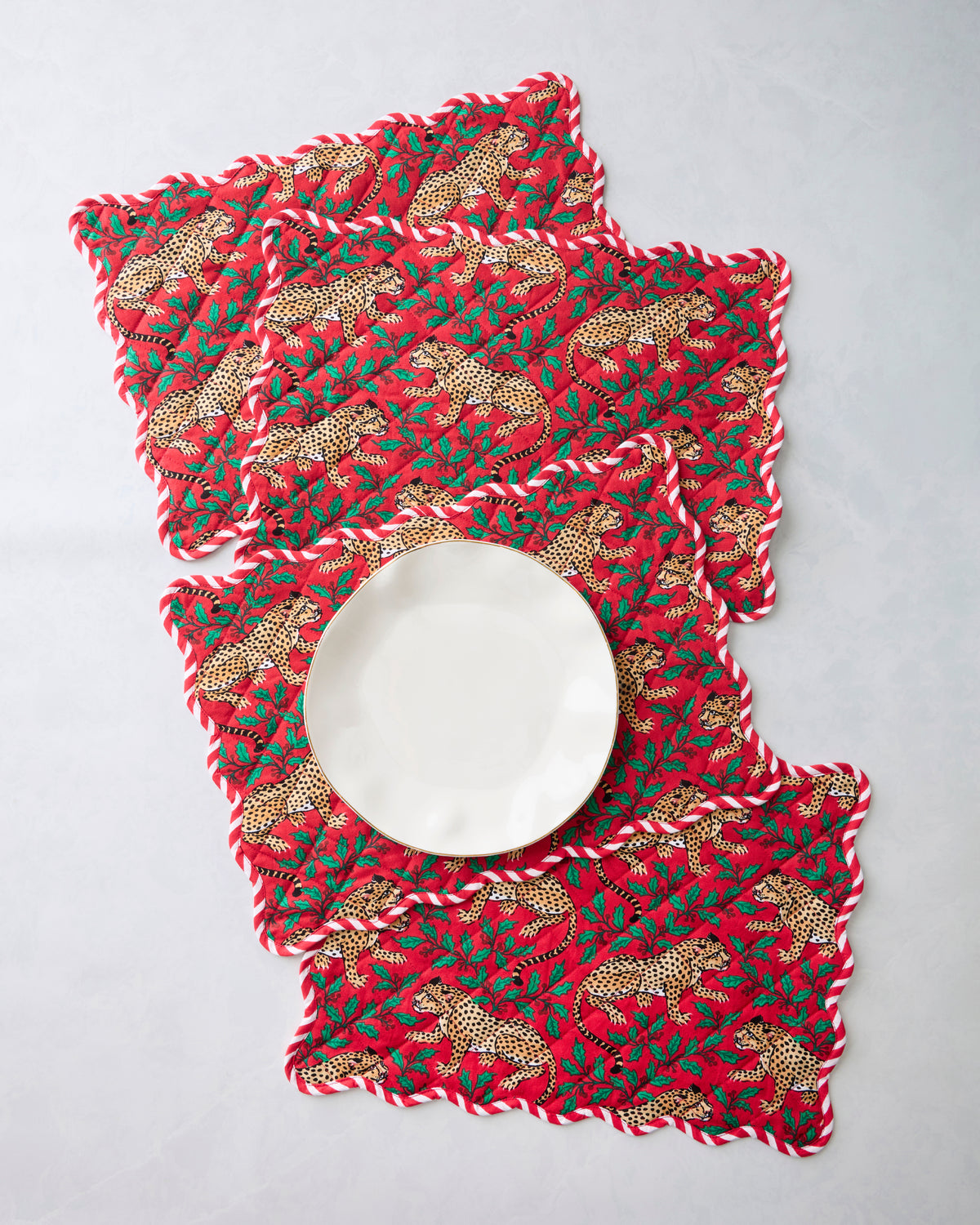 Four ruby placemats with scalloped edges and a leopard-and-holly print arranged around a white plate.