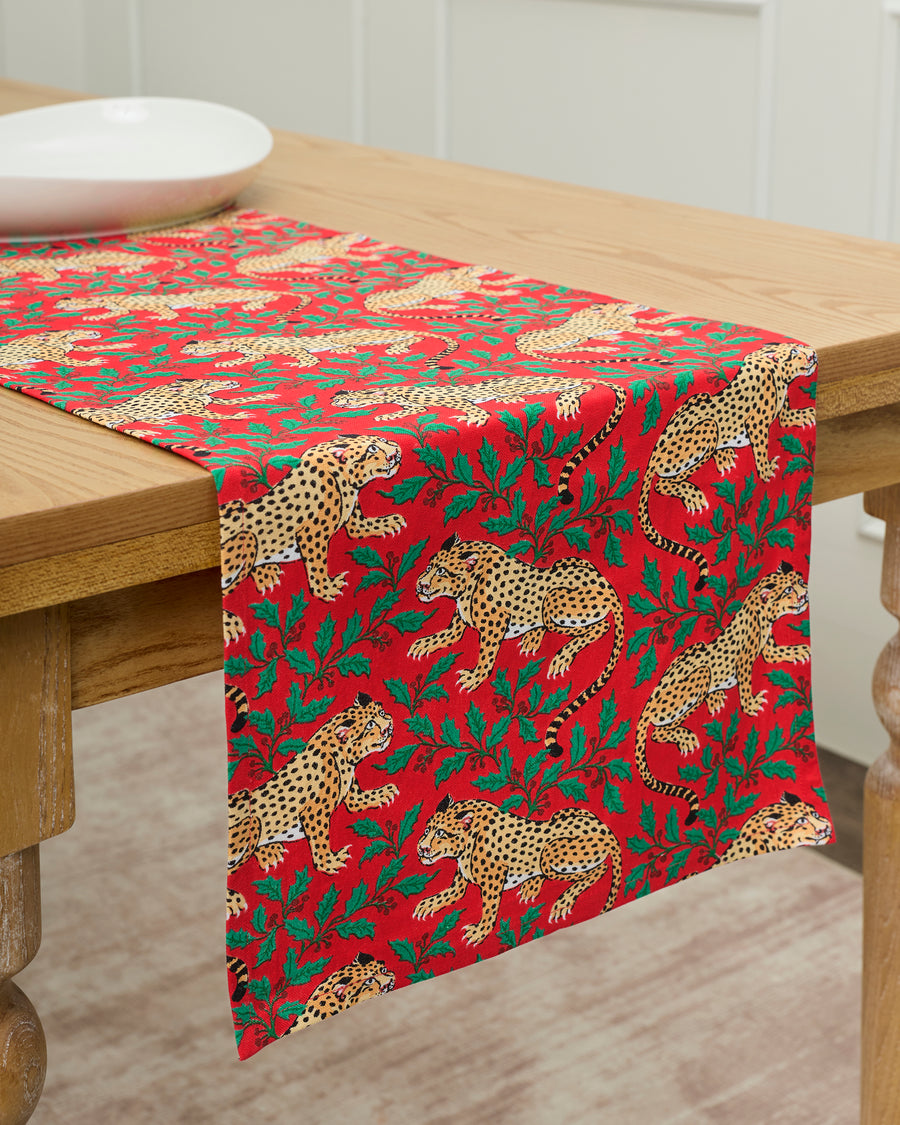 Red Winter Holly Bagheera table runner draped over a wooden table, printed with spotted leopards and holly leaves.