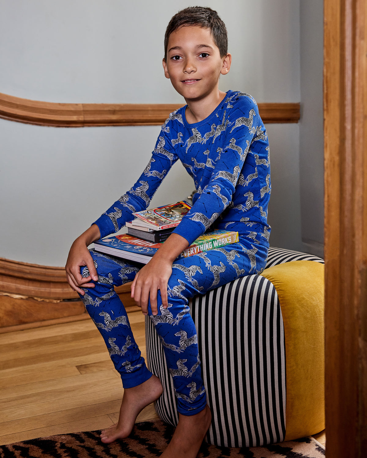 Child wearing a blue long-sleeve pajama set covered in zebra prints, seated with books on a striped chair.