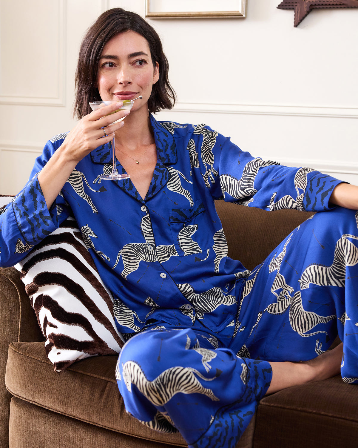 Person seated on a sofa wearing a blue zebra-print long-sleeve pajama shirt and pants, holding a cocktail glass.