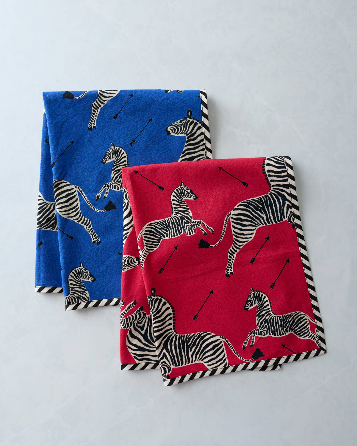 Two folded zebra-print tea towels in blue and red, each with black-and-white striped edging