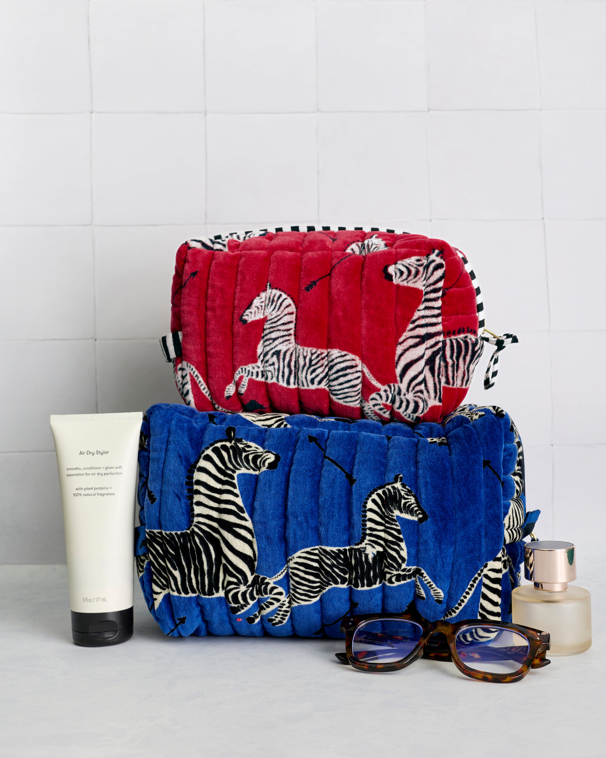 Red and blue zebra-print quilted pouches stacked with skincare, fragrance, and sunglasses.
