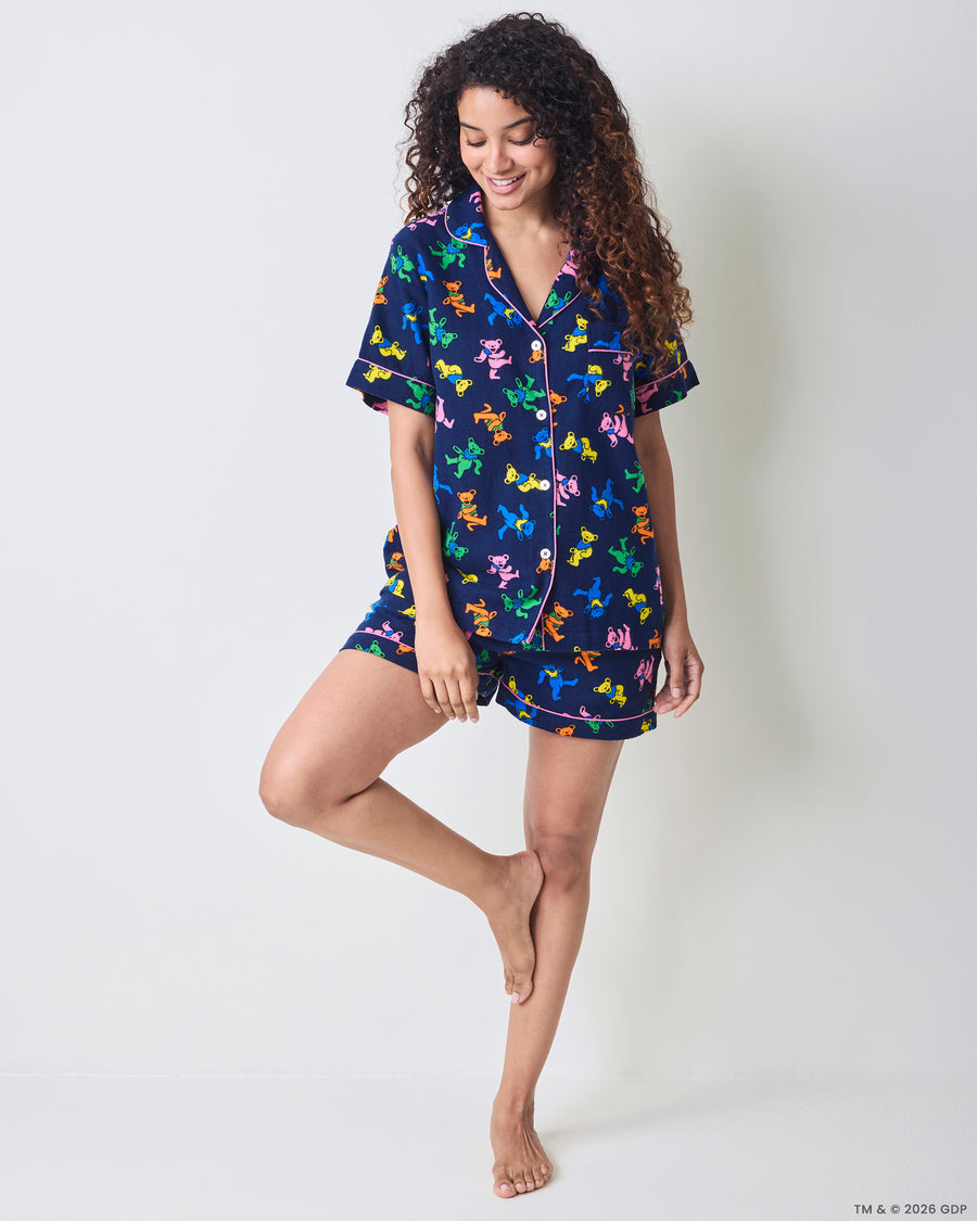Full-body view of a person wearing a navy short pajama set with a multicolor dancing-bear print, button-front top, and matching shorts.