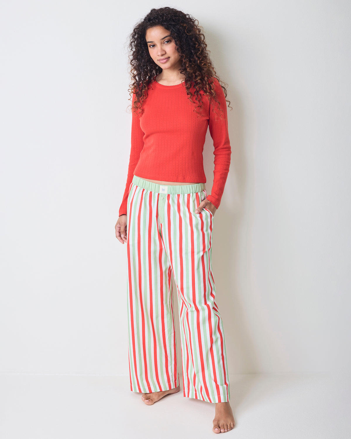 Model wearing Elf Stripes Nola pajama pants with vertical sage and cherry stripes, shown full length with a red long-sleeved top.