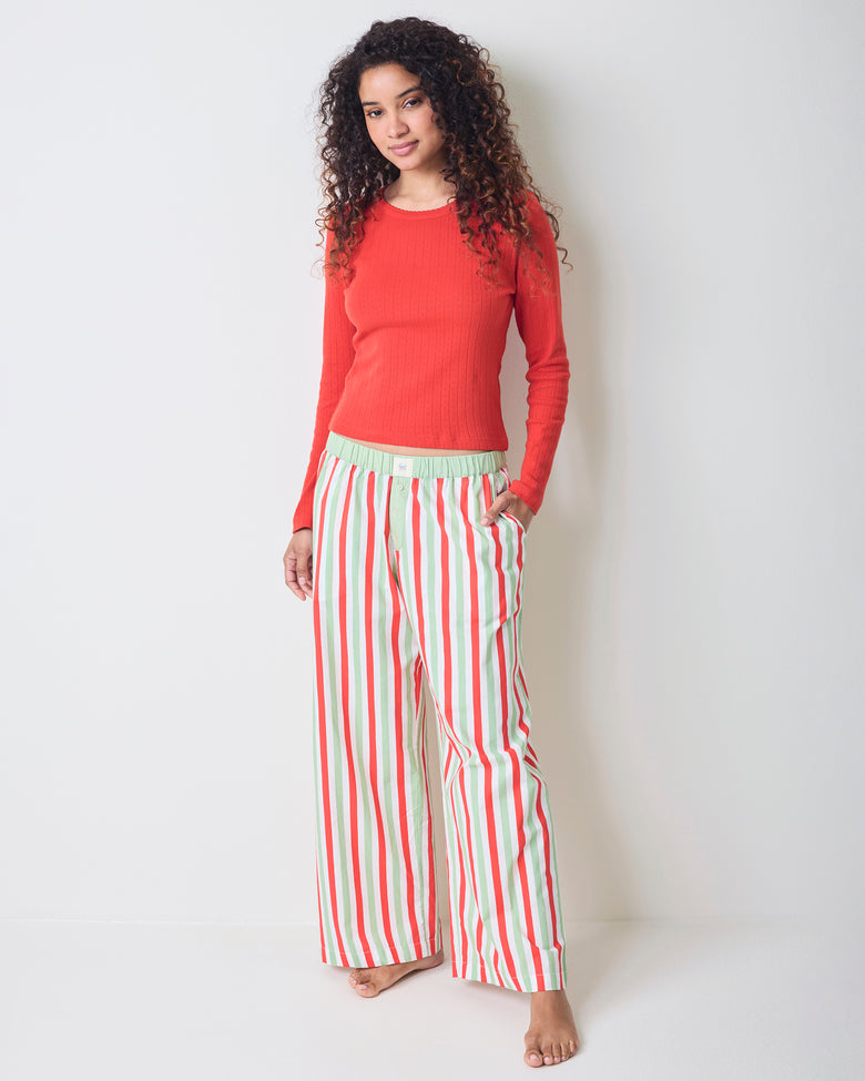 Model wearing Elf Stripes Nola pajama pants with vertical sage and cherry stripes, shown full length with a red long-sleeved top.