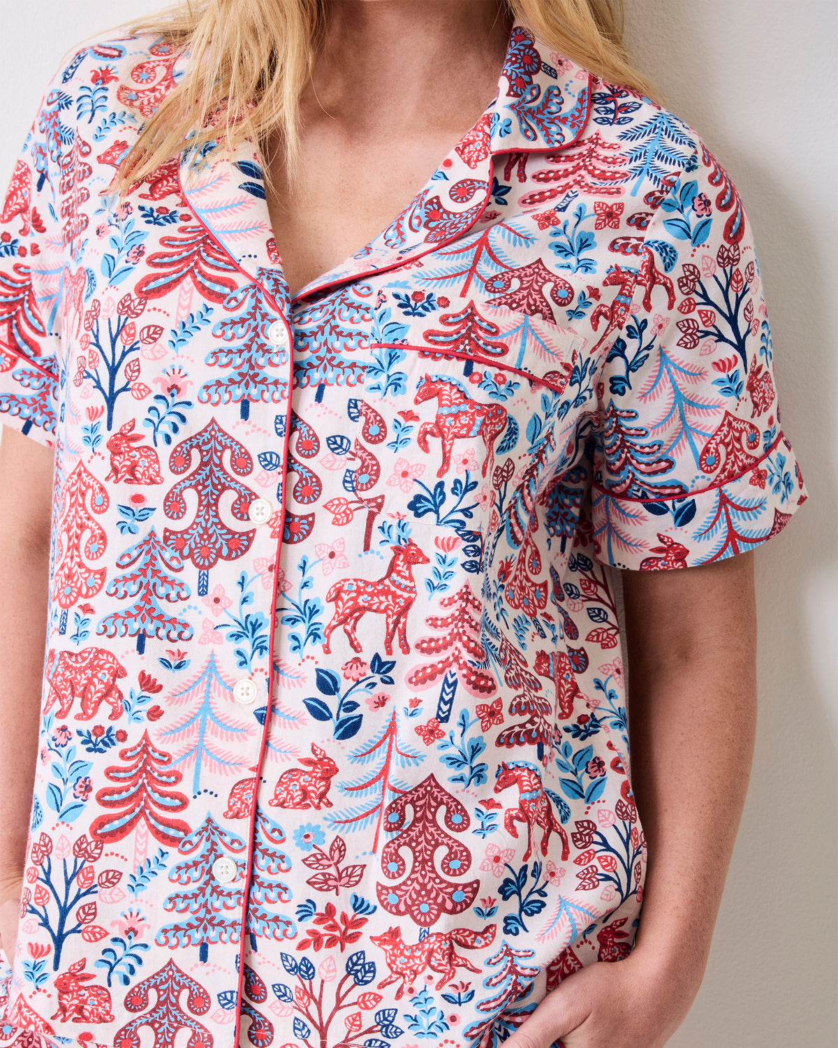 Close view of the cream short-sleeve pajama top with a collared neckline, buttons, chest pocket, and red and blue forest-inspired print.