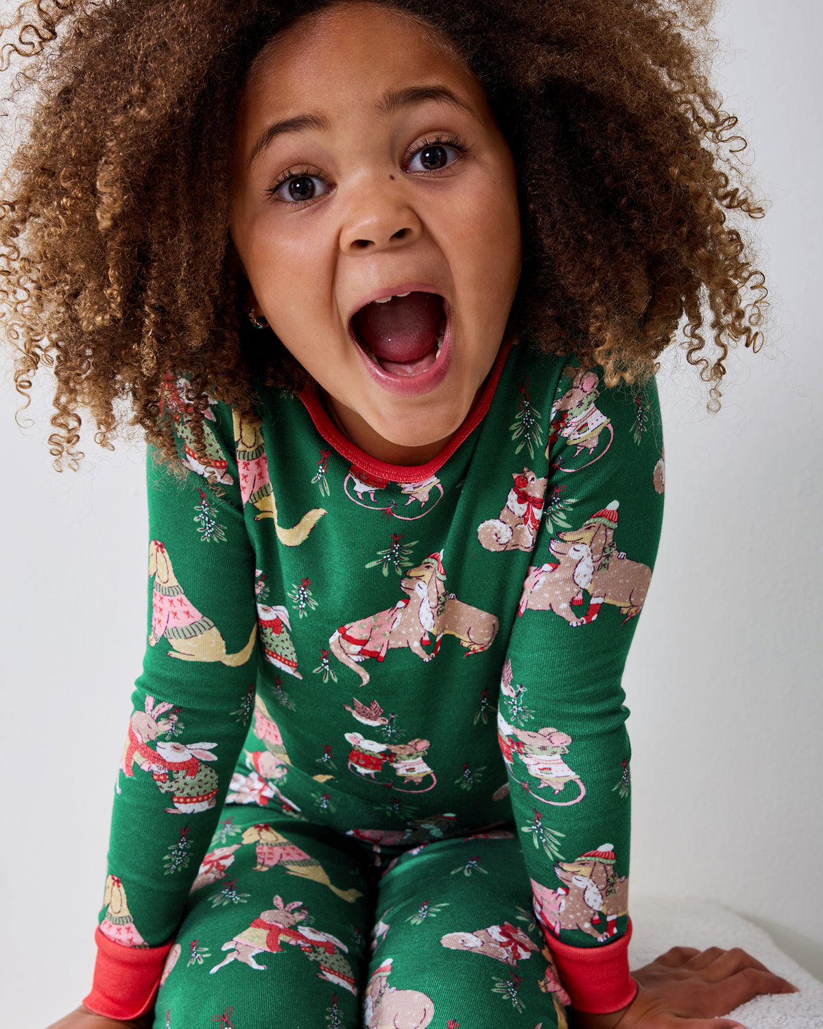 Child model wearing a green long-sleeve pajama set with red cuffs and a repeating holiday animal print.