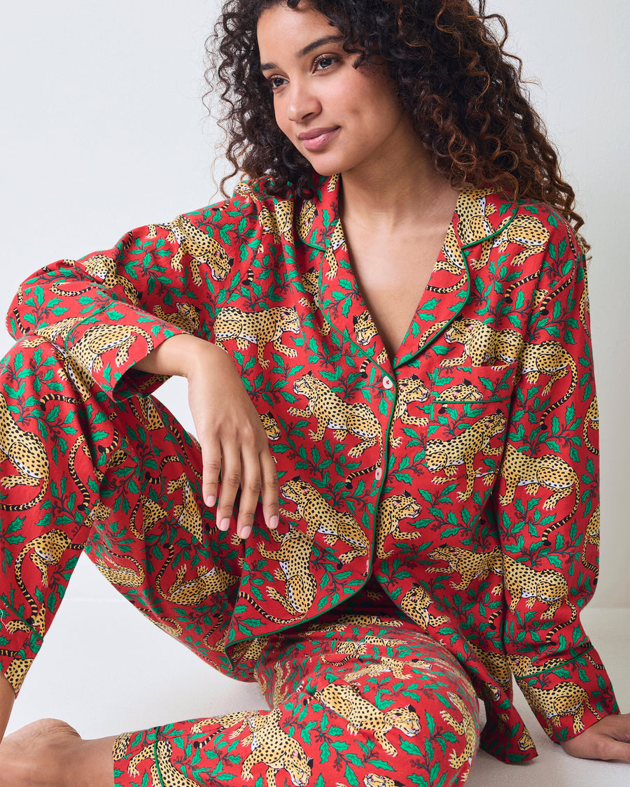 Seated person wearing the red Winter Holly Bagheera pajama top and matching pants, showing the leopard-and-holly print.