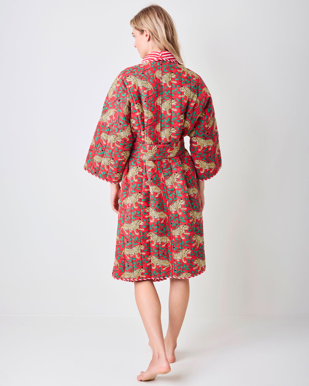 Back view of a red reversible quilted robe with a leopard and green holly print, matching belt, and striped collar trim.