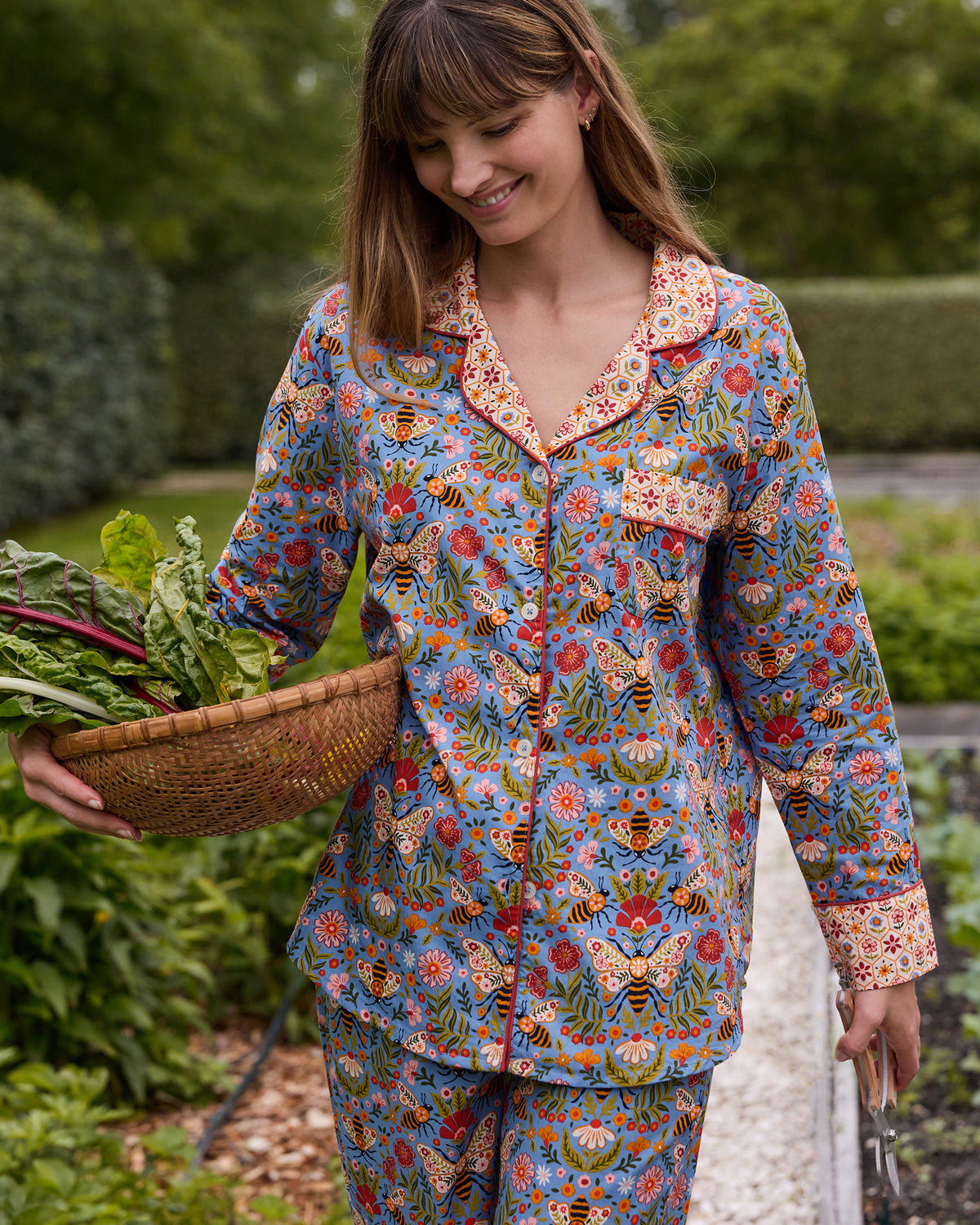 Person wearing the Delphinium Blue Bee Tapestry long pajama set and carrying a woven basket of leafy vegetables in a garden.