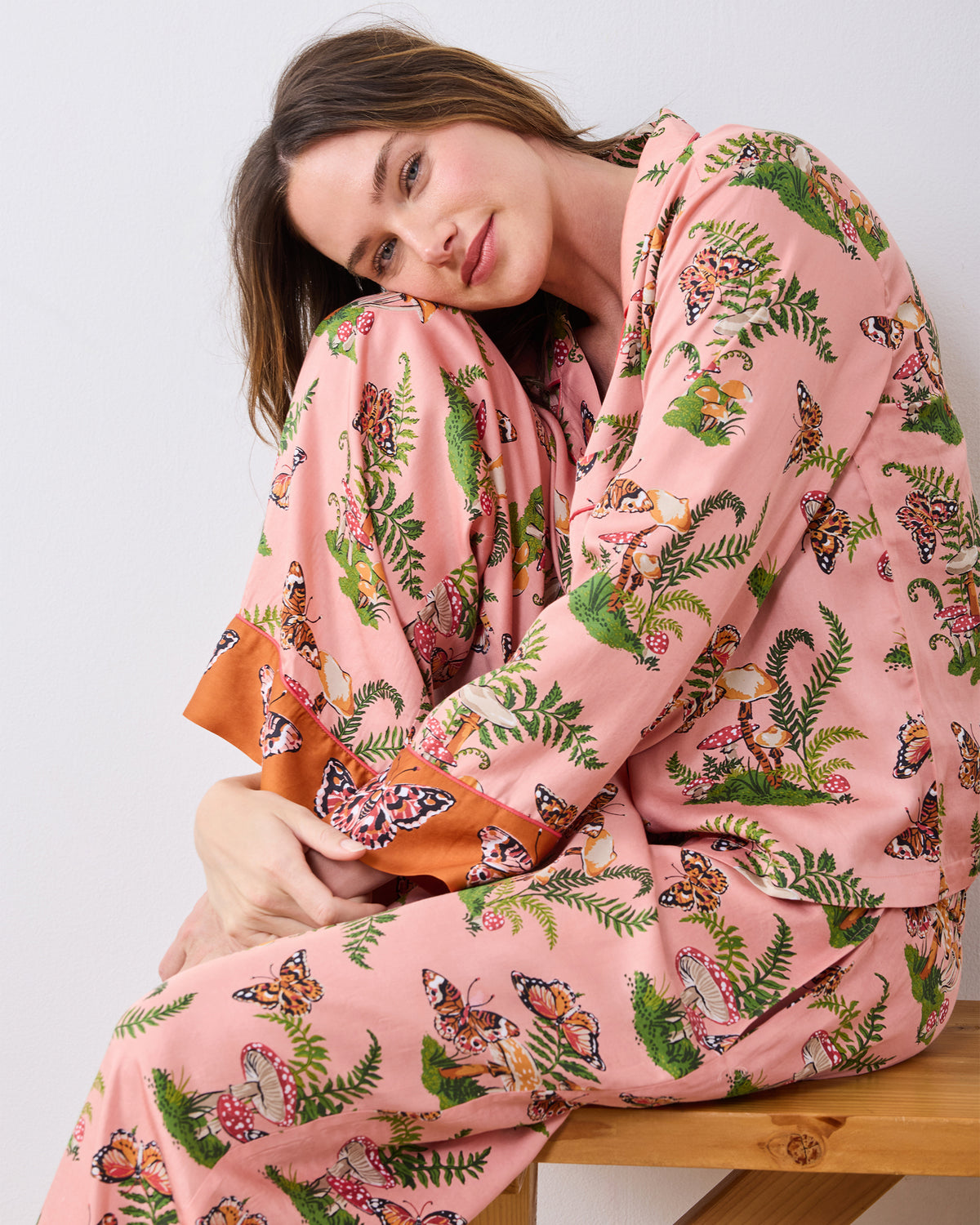 Close view of a model seated on a wooden bench wearing the pink Butterfly Forager tall pajama set, showing the patterned top, pants, and contrasting cuffs.