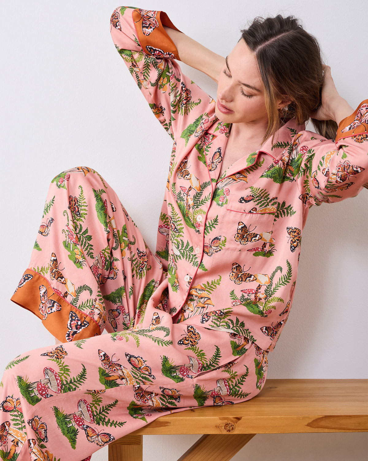Person wearing the pink Butterfly Forager tall satin Soho pajama set with long sleeves, wide-leg pants, and contrasting orange cuffs, seated on a wooden bench.