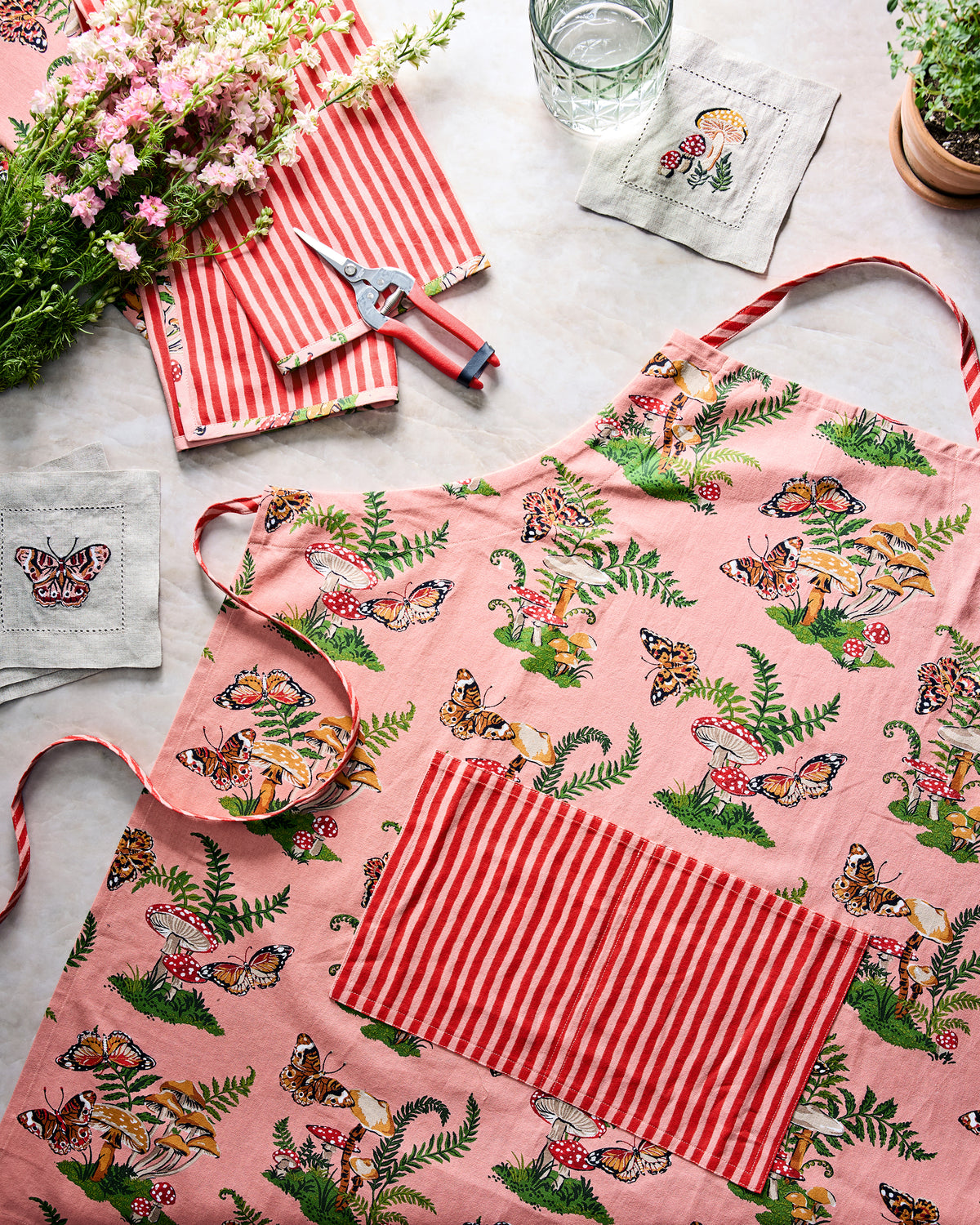 Flat lay of the rosewood Butterfly Forager apron with a front pocket, surrounded by flowers, pruning shears, striped cloth, embroidered napkins, and a glass.