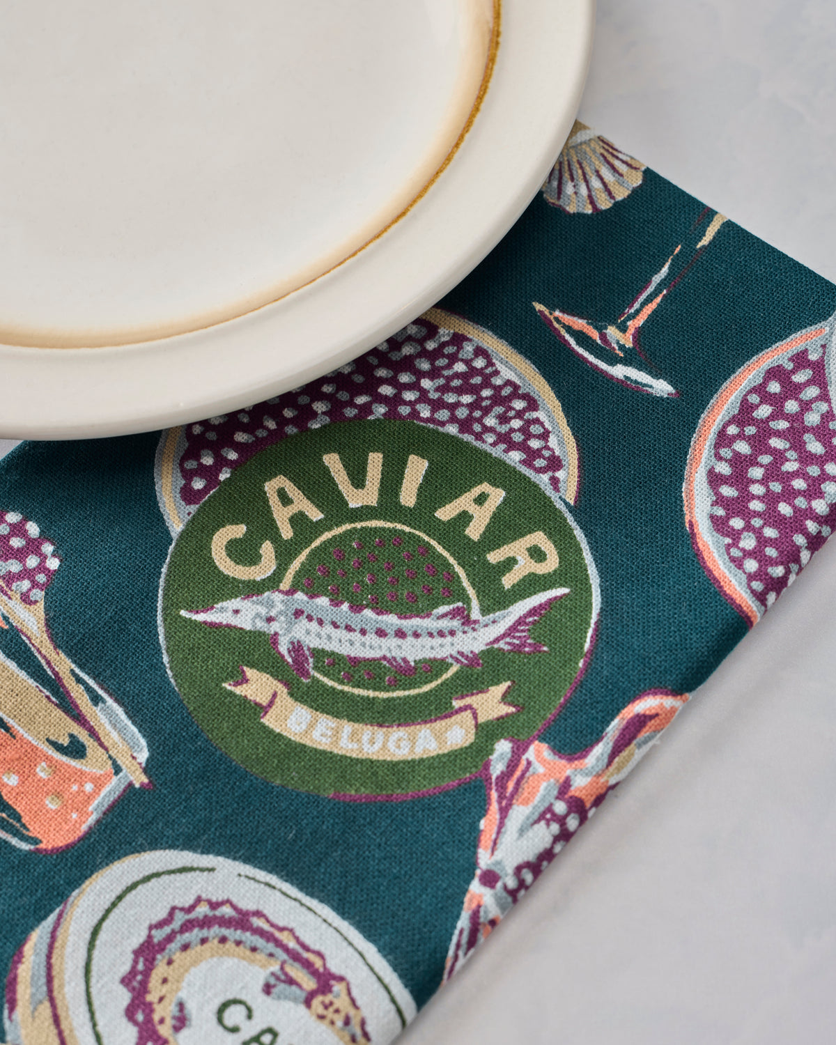 Close-up of a Champagne & Caviar dinner napkin with a green caviar emblem, shown beneath a white plate.