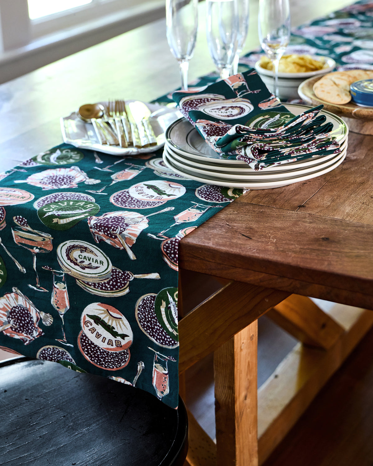 Champagne & Caviar table runner placed lengthwise on a wooden dining table beside stacked plates and folded printed napkins.