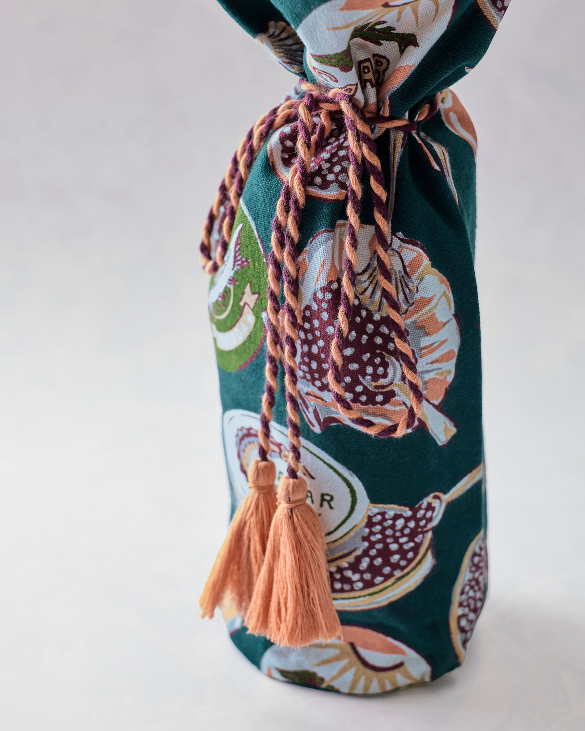 Close-up of the Champagne and Caviar wine bag with braided wrap-around tie, peach tassels, and dark teal illustrated pattern.
