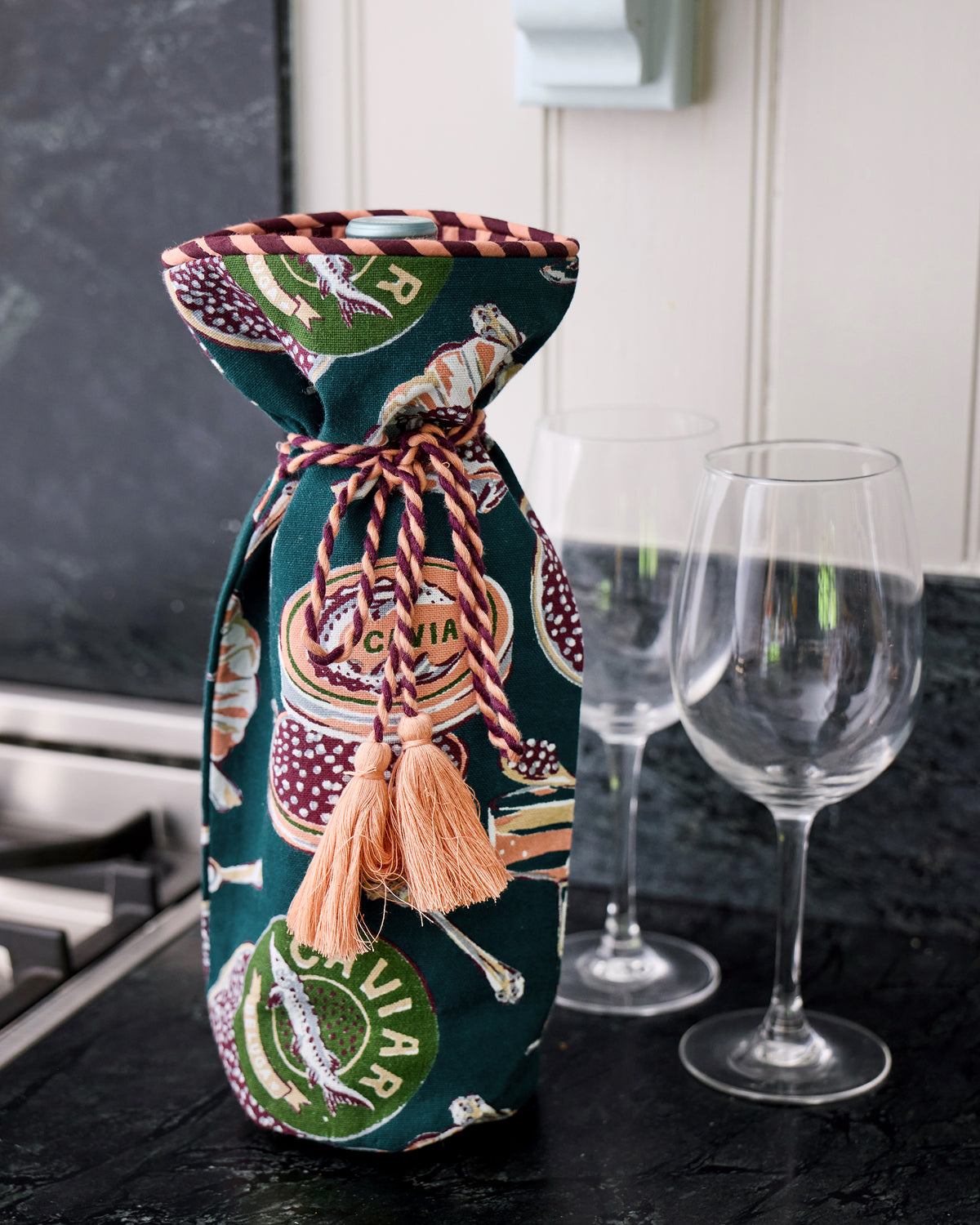 Champagne and Caviar wine bag standing on a kitchen counter beside two wine glasses, with braided ties and peach tassels.