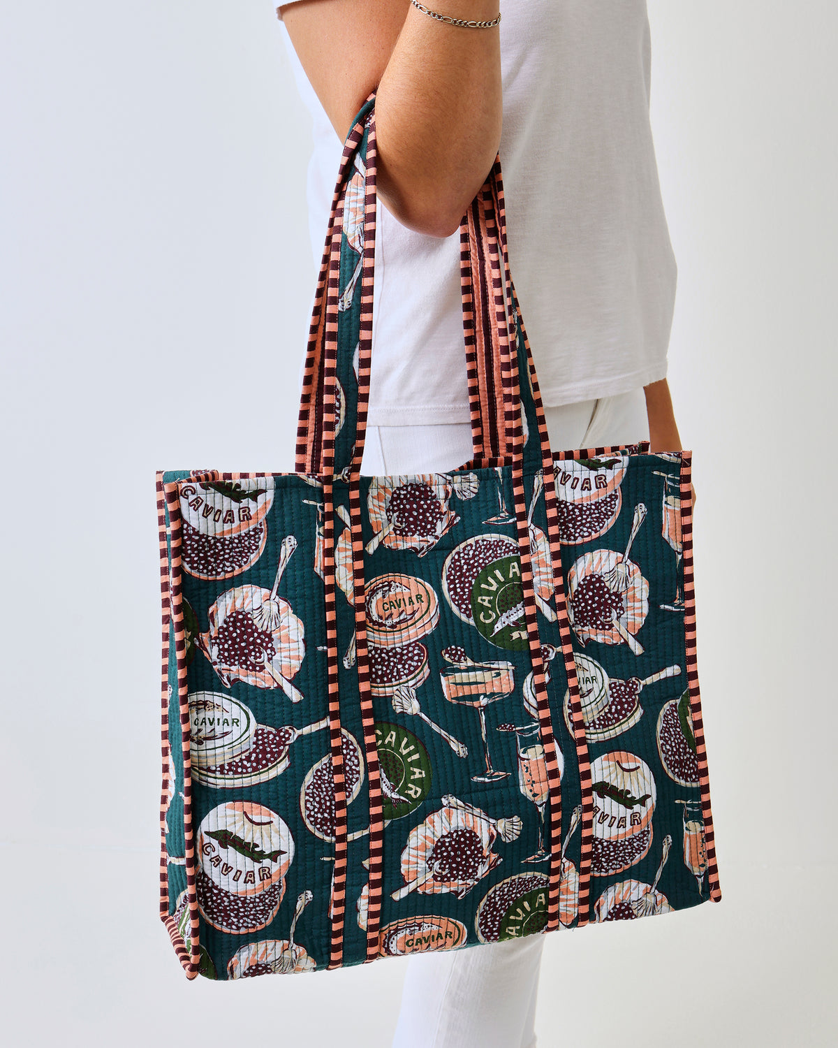 Side and back view of the Champagne & Caviar Traveler Tote with peacock-feather print, carried over a person’s shoulder.