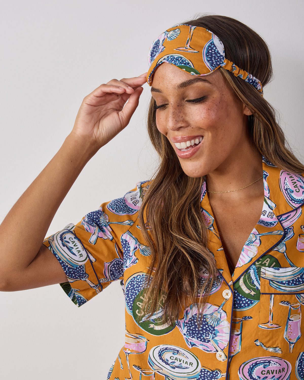 Goldenrod Champagne & Caviar eye mask with an adjustable strap, shown worn on a person with a matching printed pajama top.