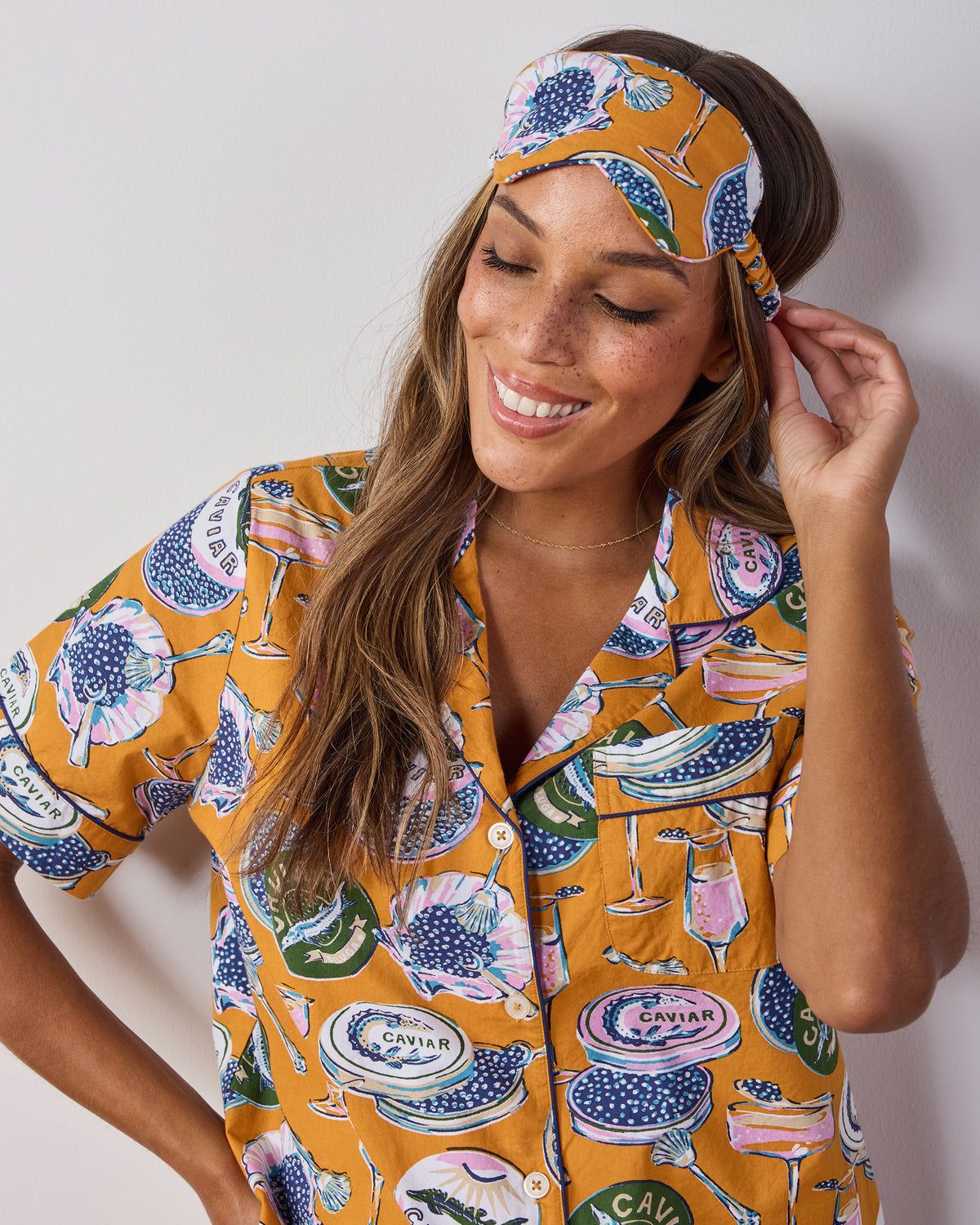Front view of a Goldenrod Champagne & Caviar eye mask with an adjustable strap, shown worn on a person with a matching printed pajama top.