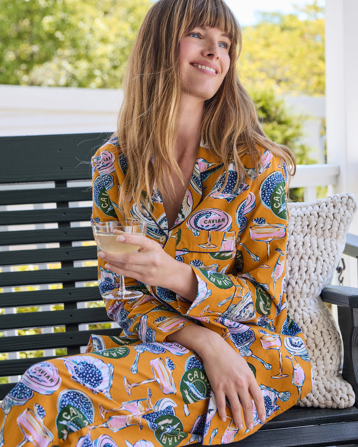 Person seated on a porch wearing the goldenrod Champagne & Caviar petite long pajama set and holding a stemmed glass.