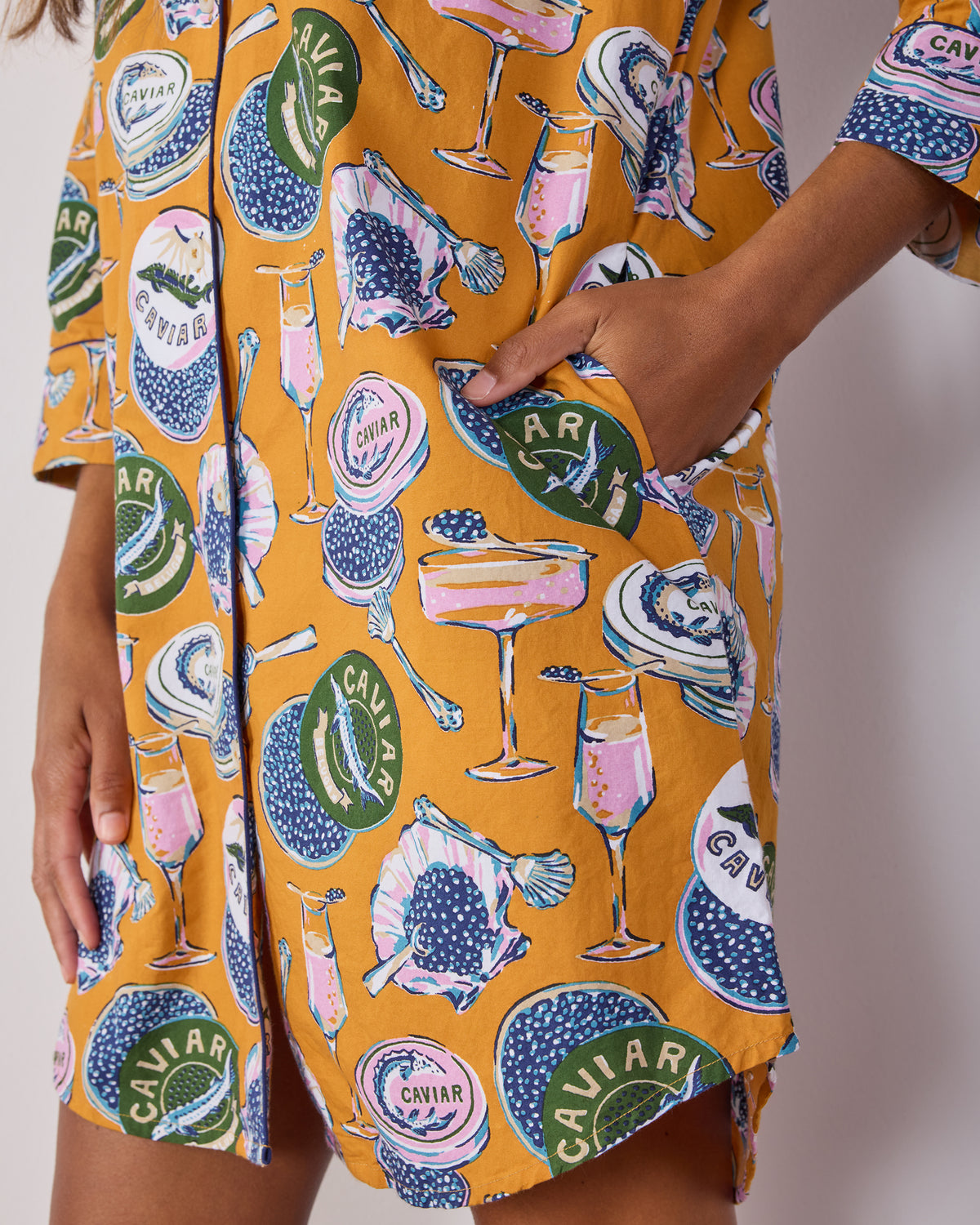 Close-up of the goldenrod Champagne & Caviar sleep shirt showing a side pocket, button placket, curved hem, and printed motifs.