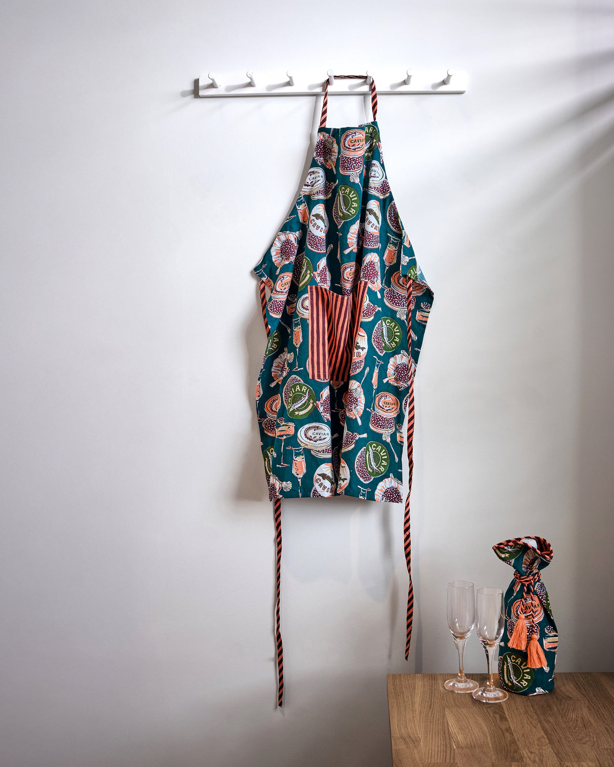 Champagne and Caviar apron hanging on a wall rack, with patterned fabric, striped neck and waist ties, and a front pocket.