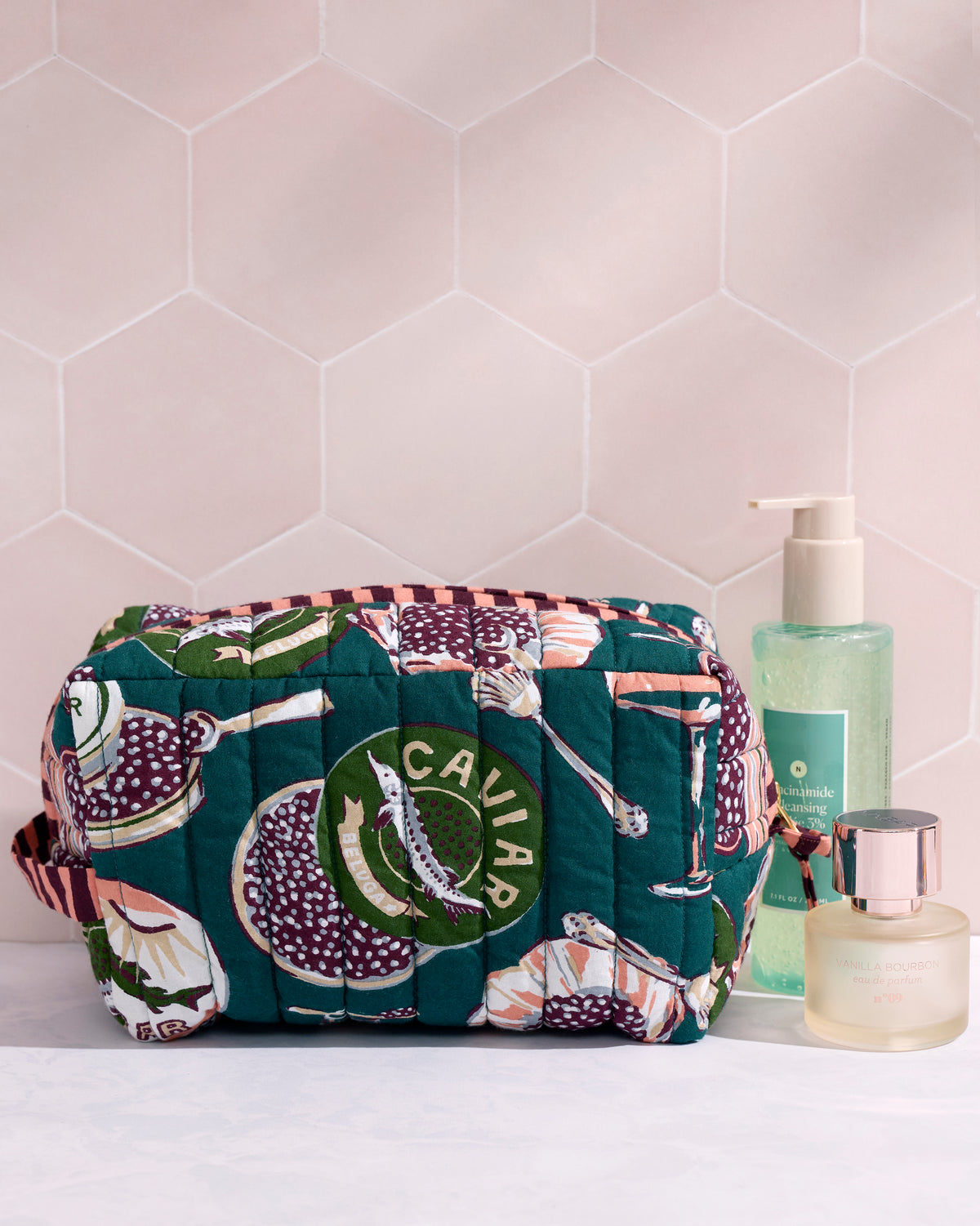 Quilted peacock-feather pouch with a Champagne & Caviar print, shown beside a pump bottle and perfume.