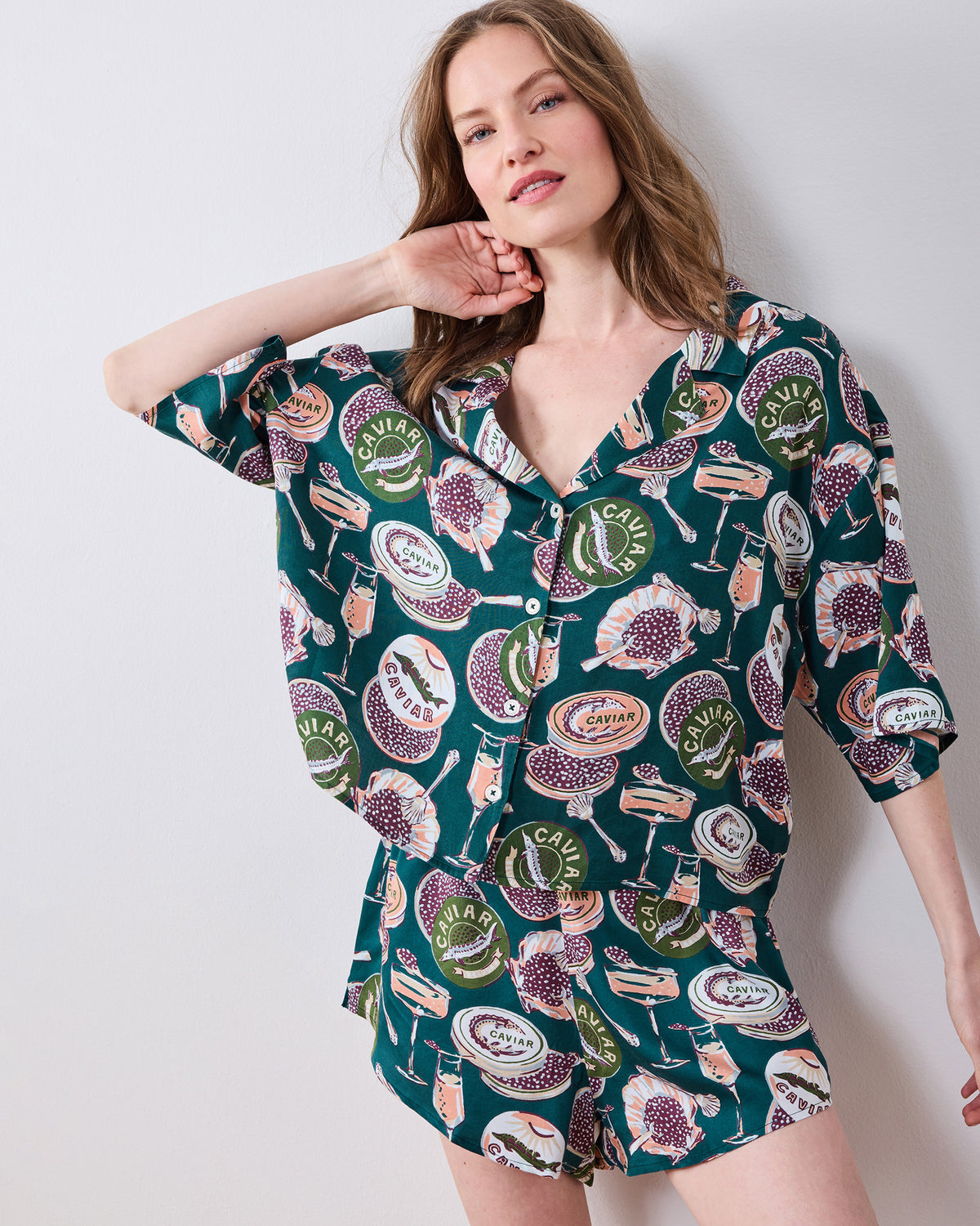 Person wearing a dark teal short pajama set with a button-front top and shorts, printed with caviar tins, champagne glasses, and spoons.