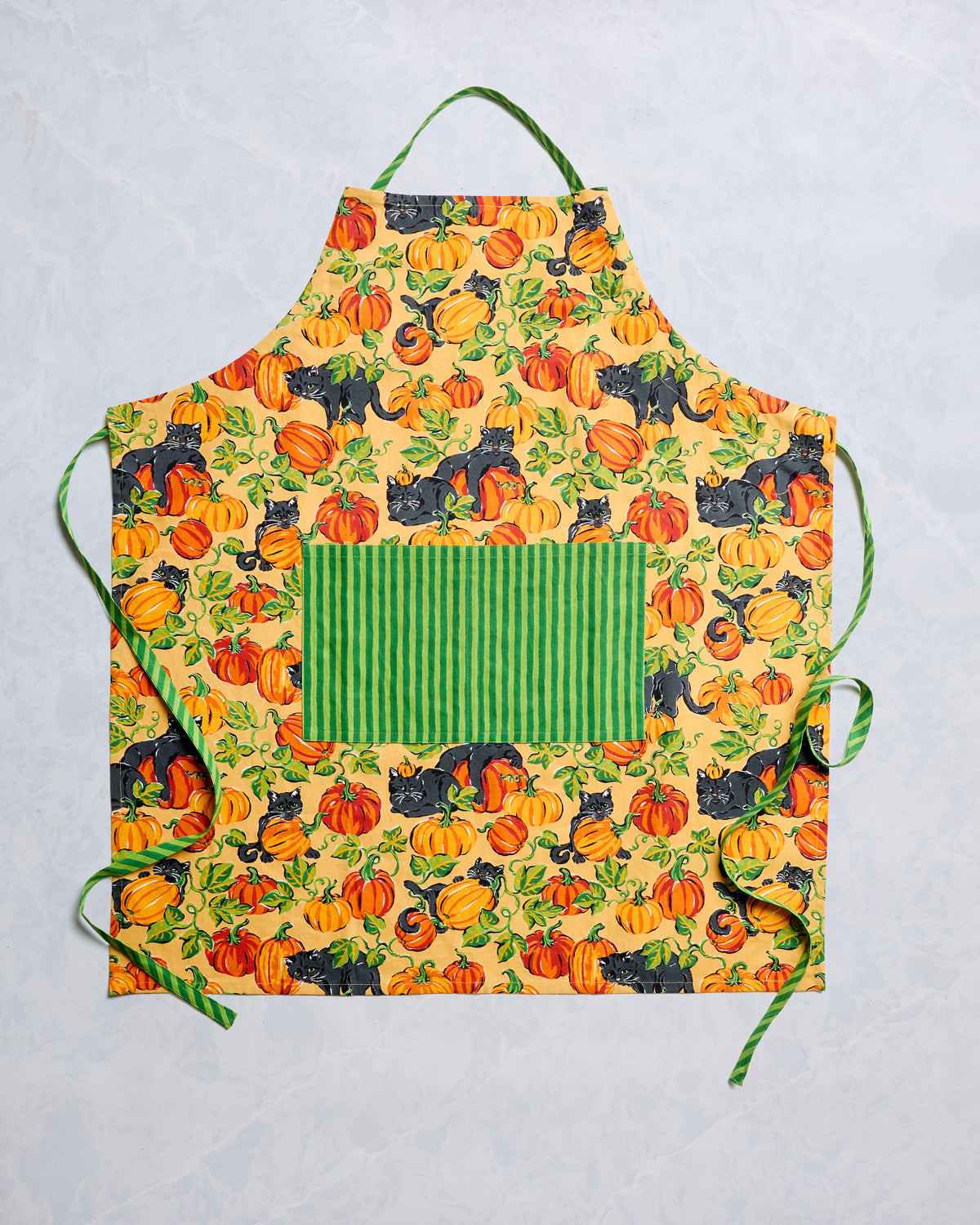 Flat lay of a pumpkin-and-black-cat print apron with green striped pocket, neck strap, and waist ties.