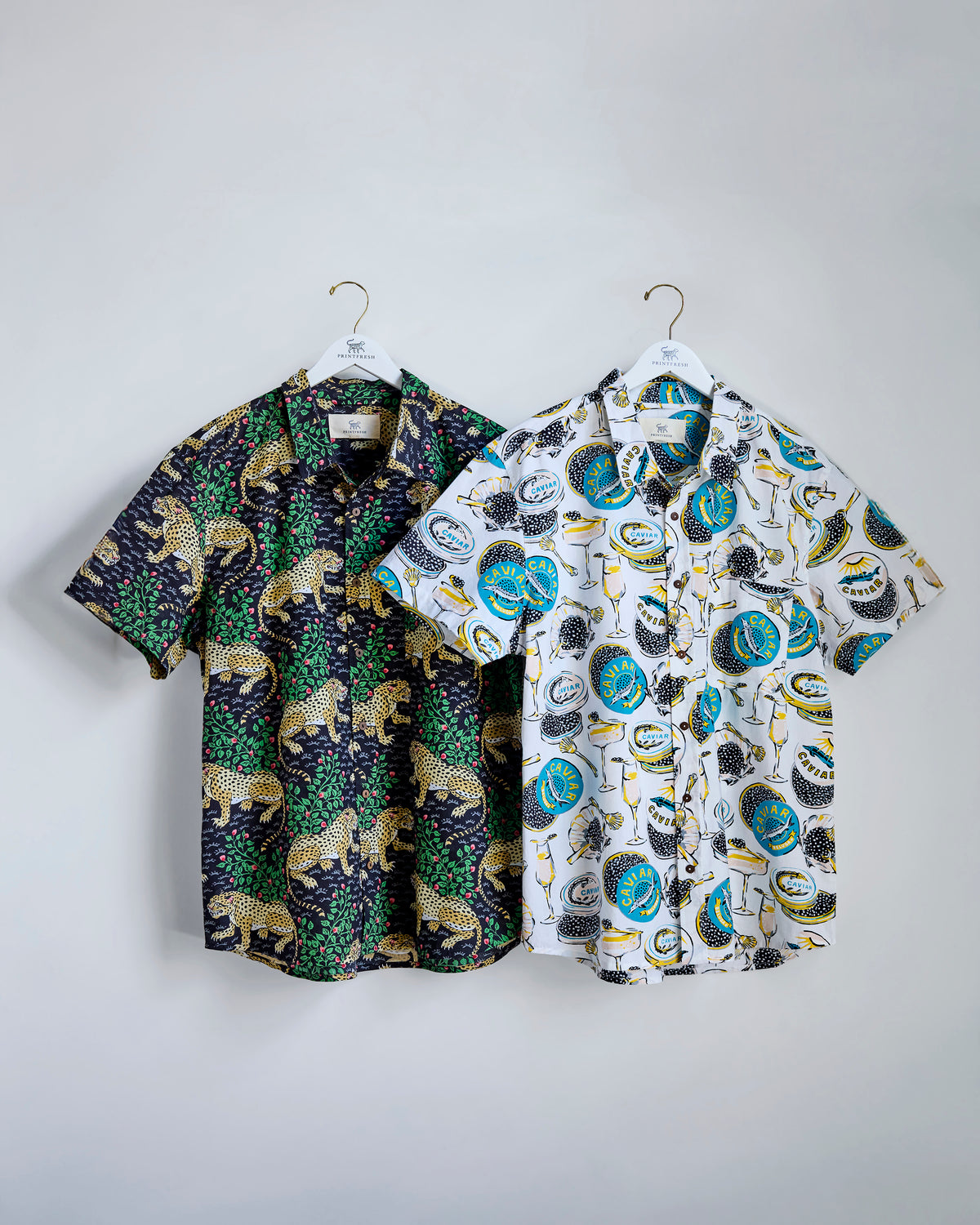 Two short-sleeve button-down shirts on hangers: one dark shirt with leopard and foliage motifs and one light shirt with a caviar-themed print.