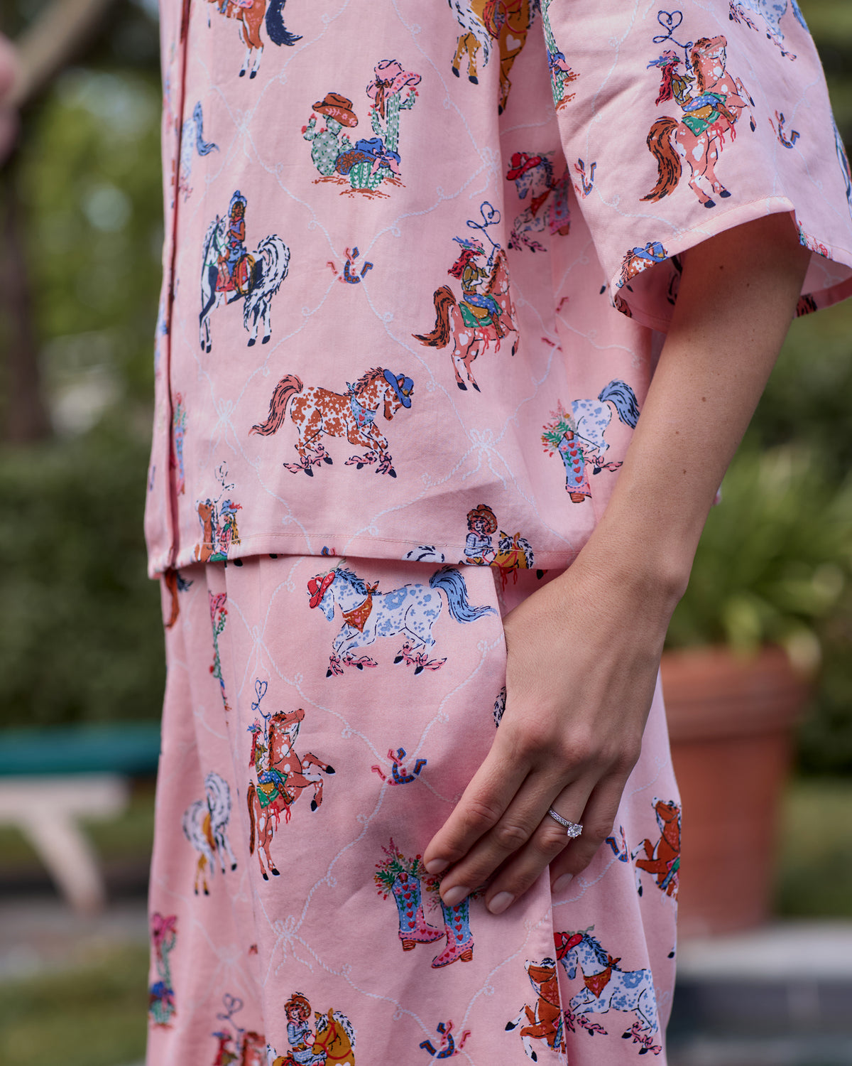 Close-up of the pink pajama top and wide-leg pants, showing the horse-and-cowgirl print and a hand resting in a pants pocket.
