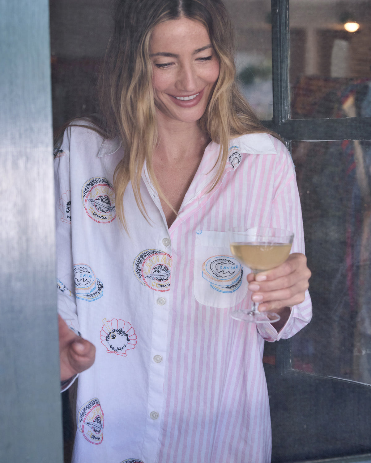Close front view of the Embroidered Caviar Club button-down sleep shirt, showing its embroidered motifs and chest pocket while holding a stemmed glass.