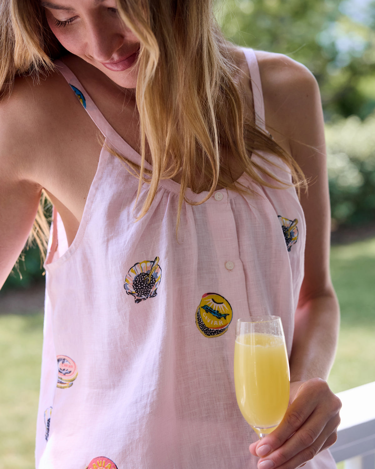 Close-up of the Pink Pearl cami top showing its shoulder straps, buttons, and embroidered caviar tin motifs beside a glass of yellow drink.