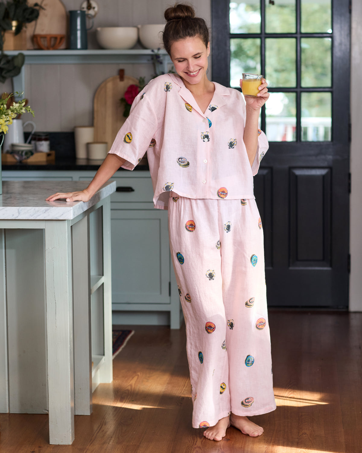 Full-length view of a person wearing the pink pearl cropped top and wide-leg pants set with colorful embroidered caviar-tin motifs, holding a glass in a kitchen.