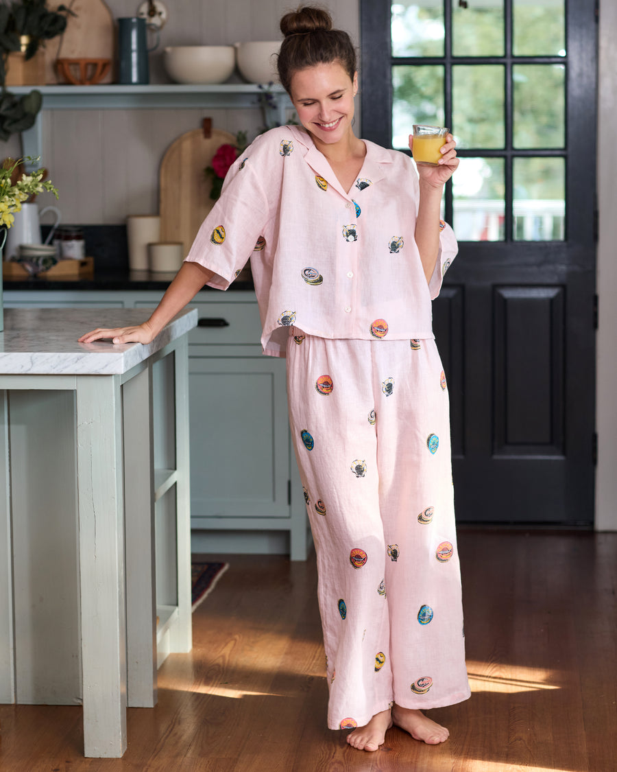 Full-length view of a person wearing the pink pearl cropped top and wide-leg pants set with colorful embroidered caviar-tin motifs, holding a glass in a kitchen.