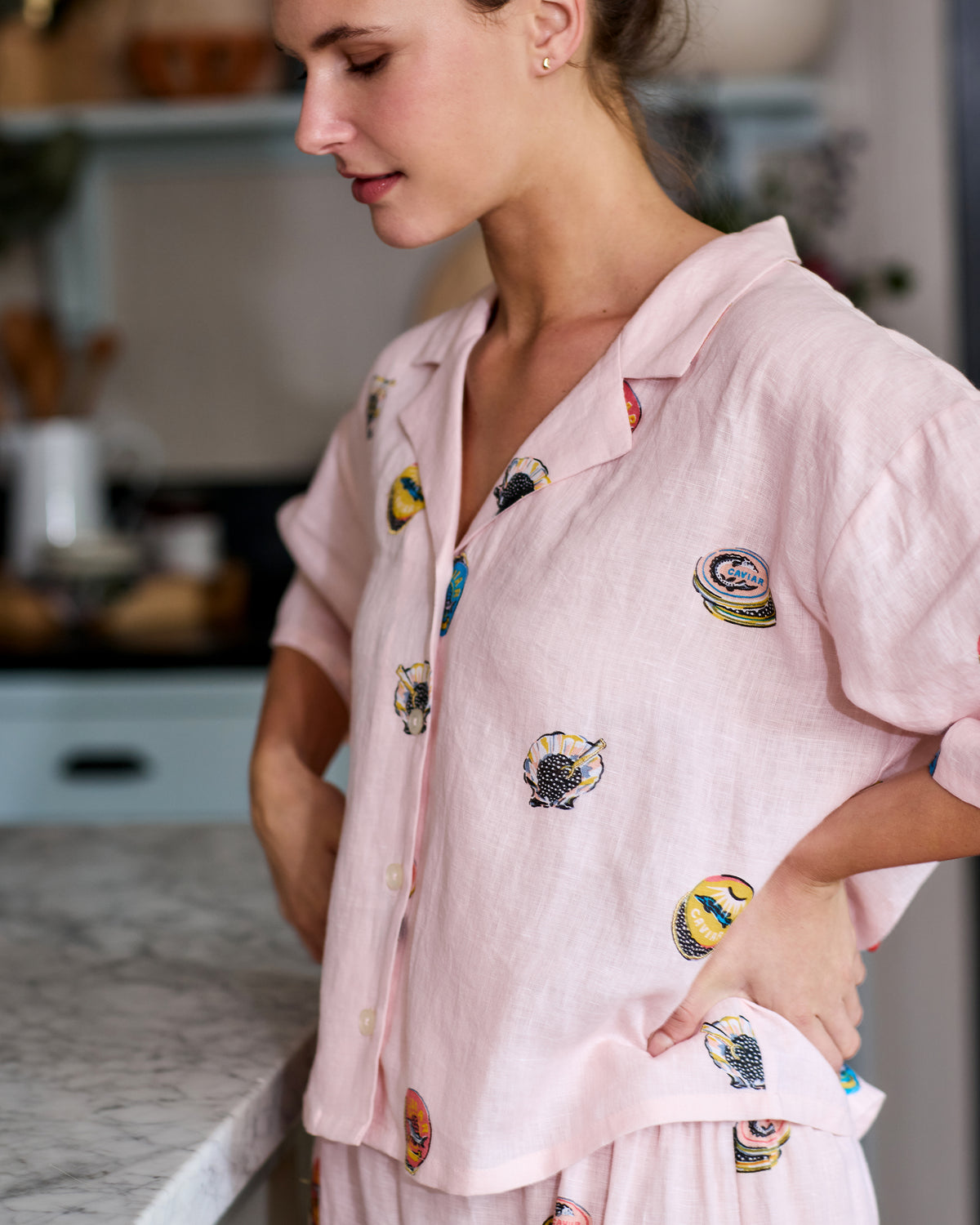 Close-up of a pink short-sleeve pajama top with embroidered caviar tins and shell motifs, shown on a model in a kitchen.