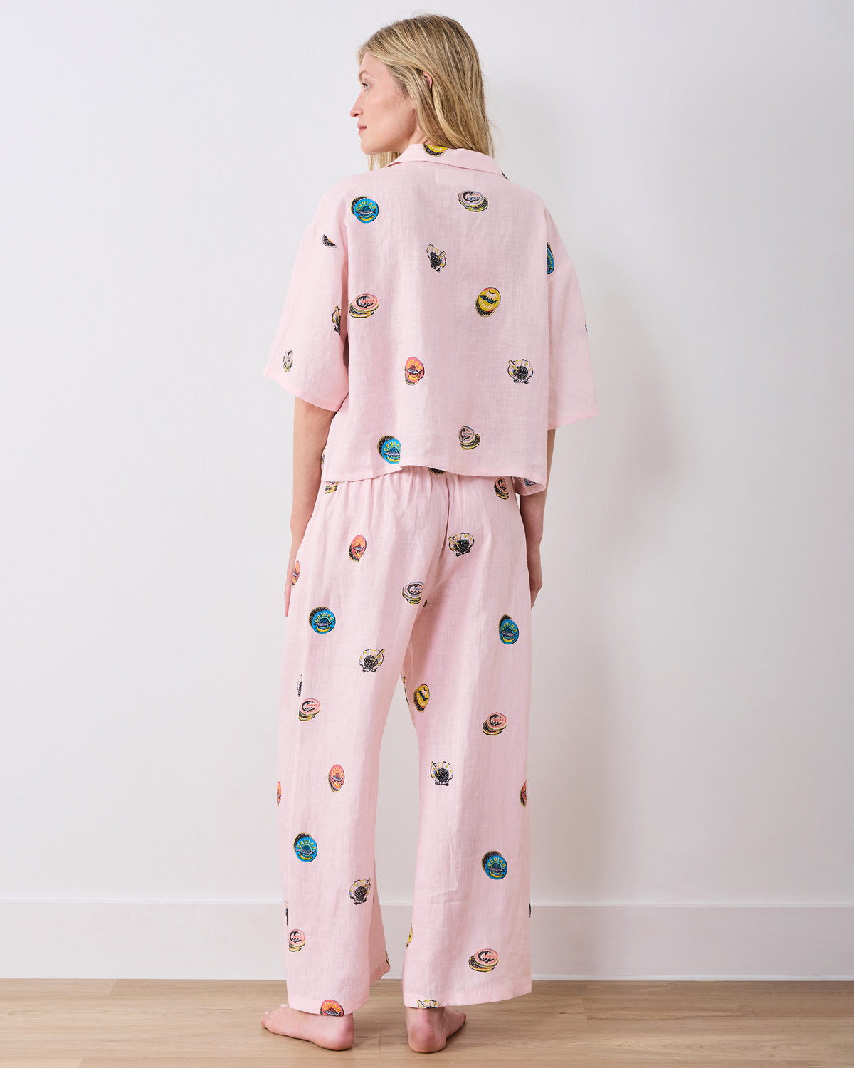 Back view of a model wearing a pink cropped top and wide-leg pants with scattered embroidered caviar tin and shell motifs.