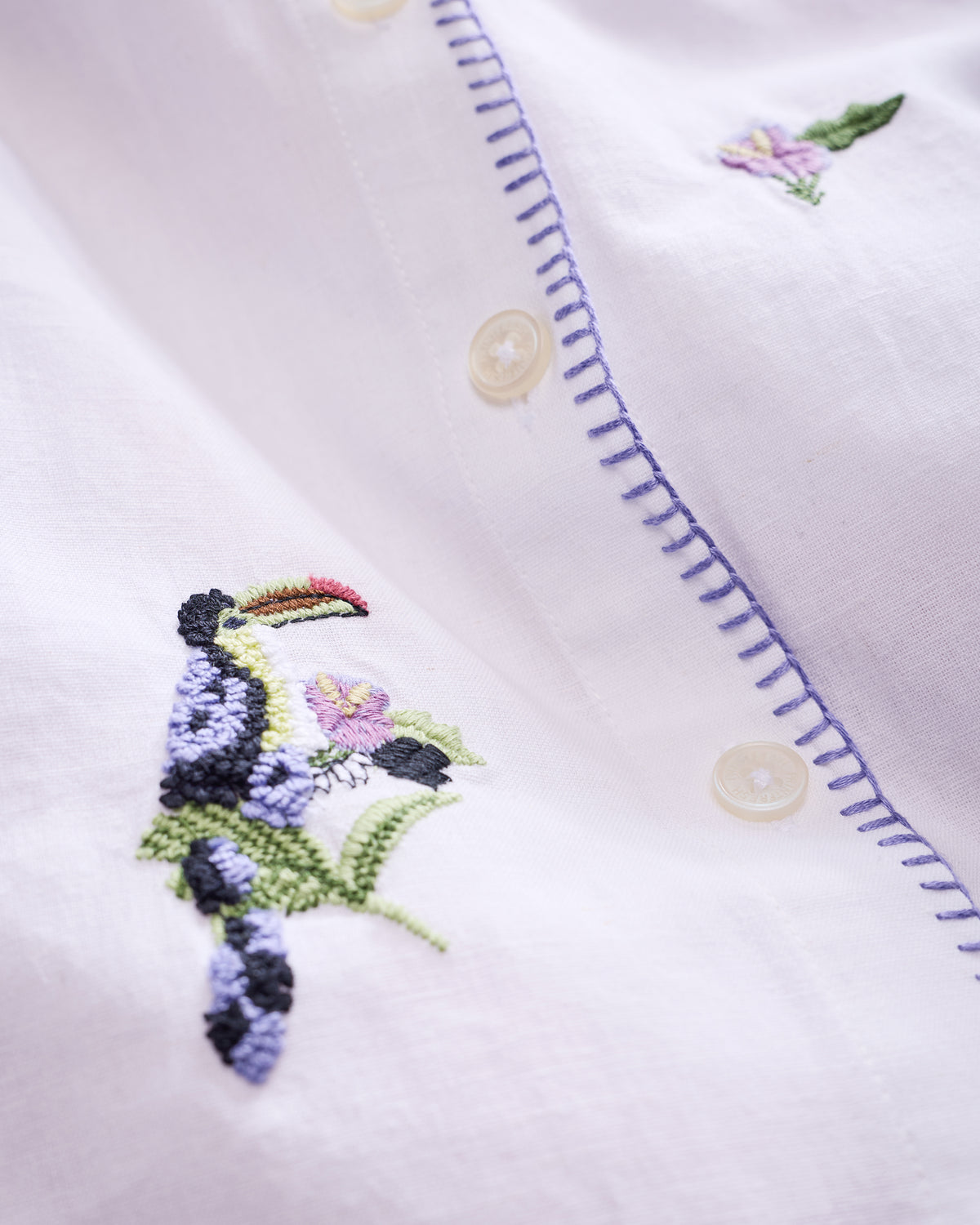 Close-up of the white button-down showing pineapple-and-toucan embroidery, cream buttons, and purple stitching along the placket.