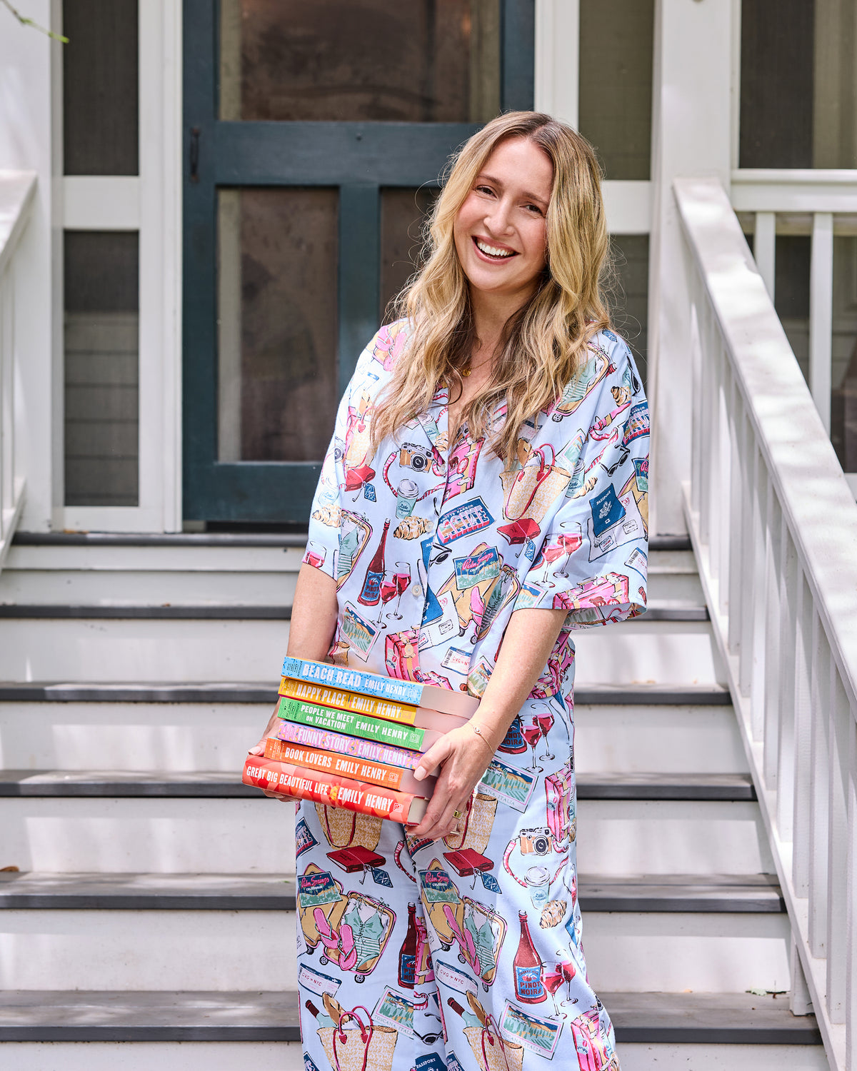 Model wearing a light blue printed short-sleeve pajama set and holding six Emily Henry novels on a porch.