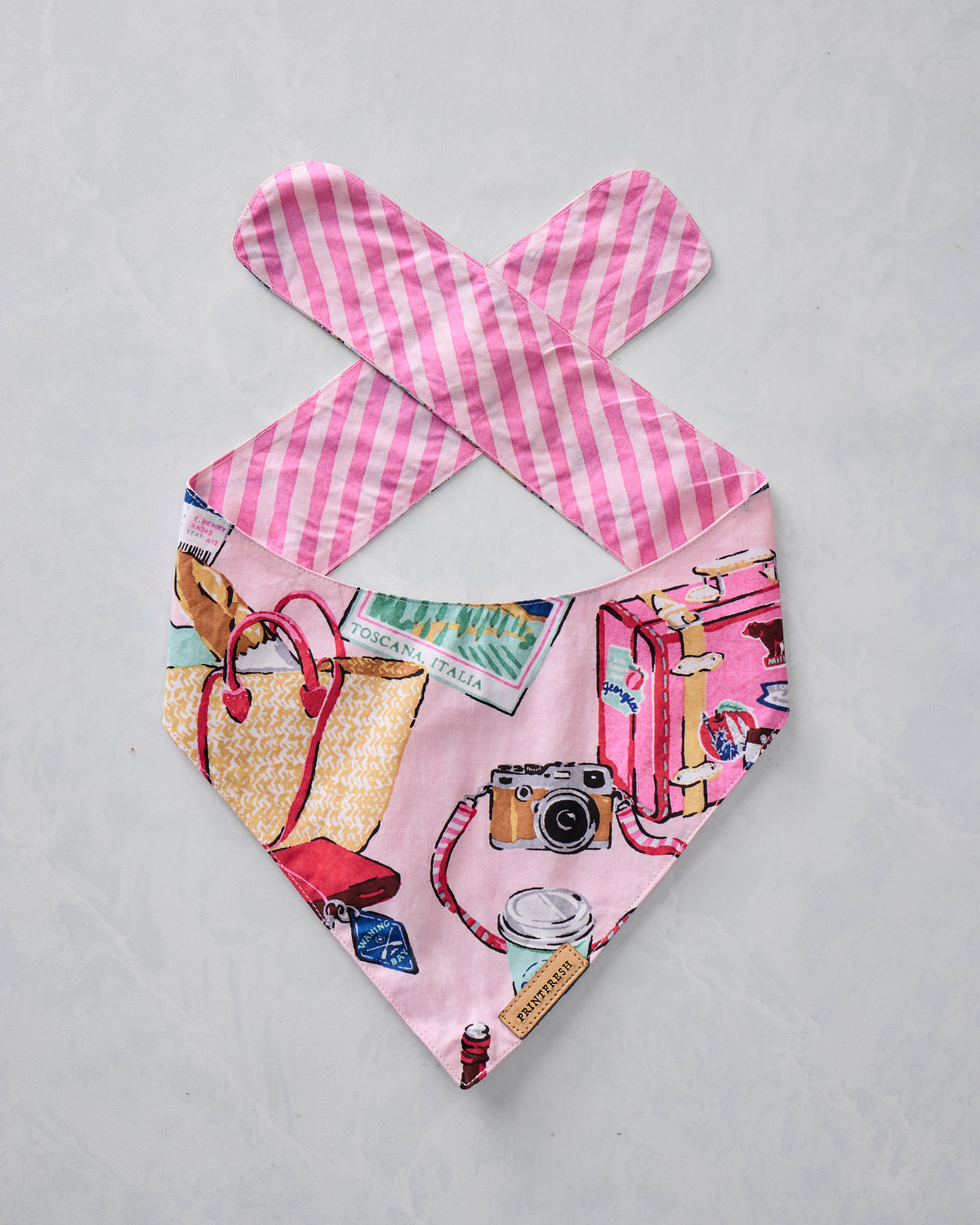 Reversible pink pet bandana with travel-themed illustrations, shown with the printed side facing up.