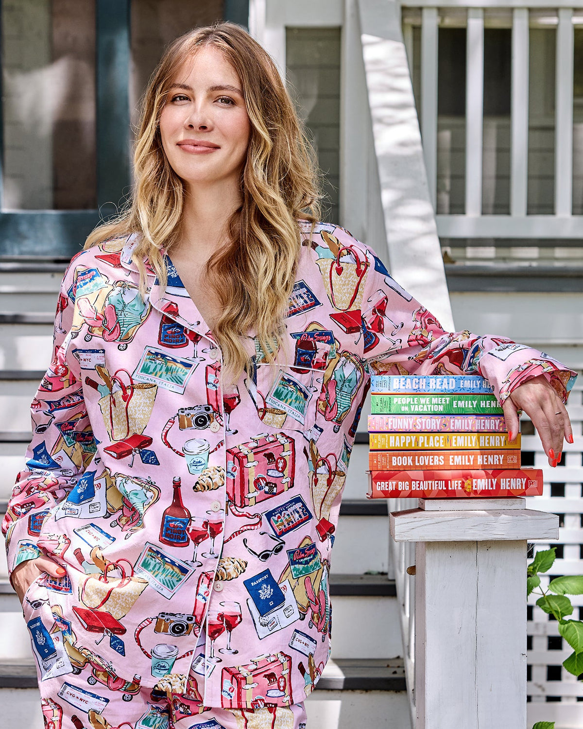Person wearing a pink illustrated button-down pajama set beside a stack of Emily Henry books outdoors.
