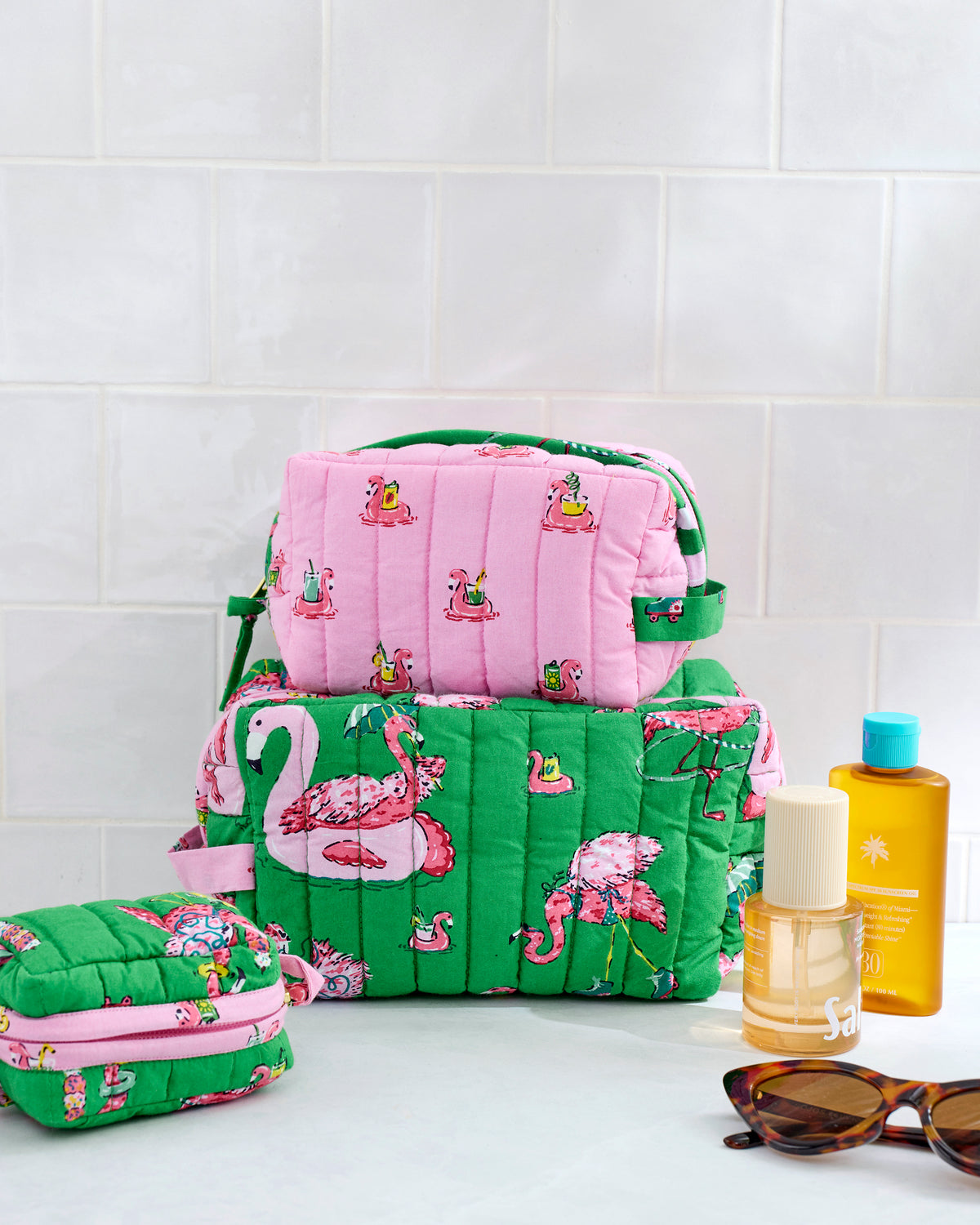 Three quilted Flock Party pouches in different sizes, with pink flamingo prints, shown with toiletries and sunglasses.