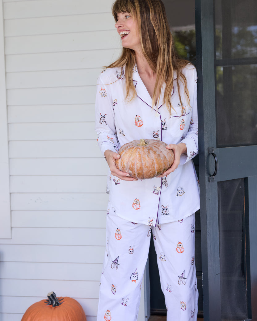 Person wearing a white long-sleeve pajama set with dog faces in costumes, holding a pumpkin in a doorway.